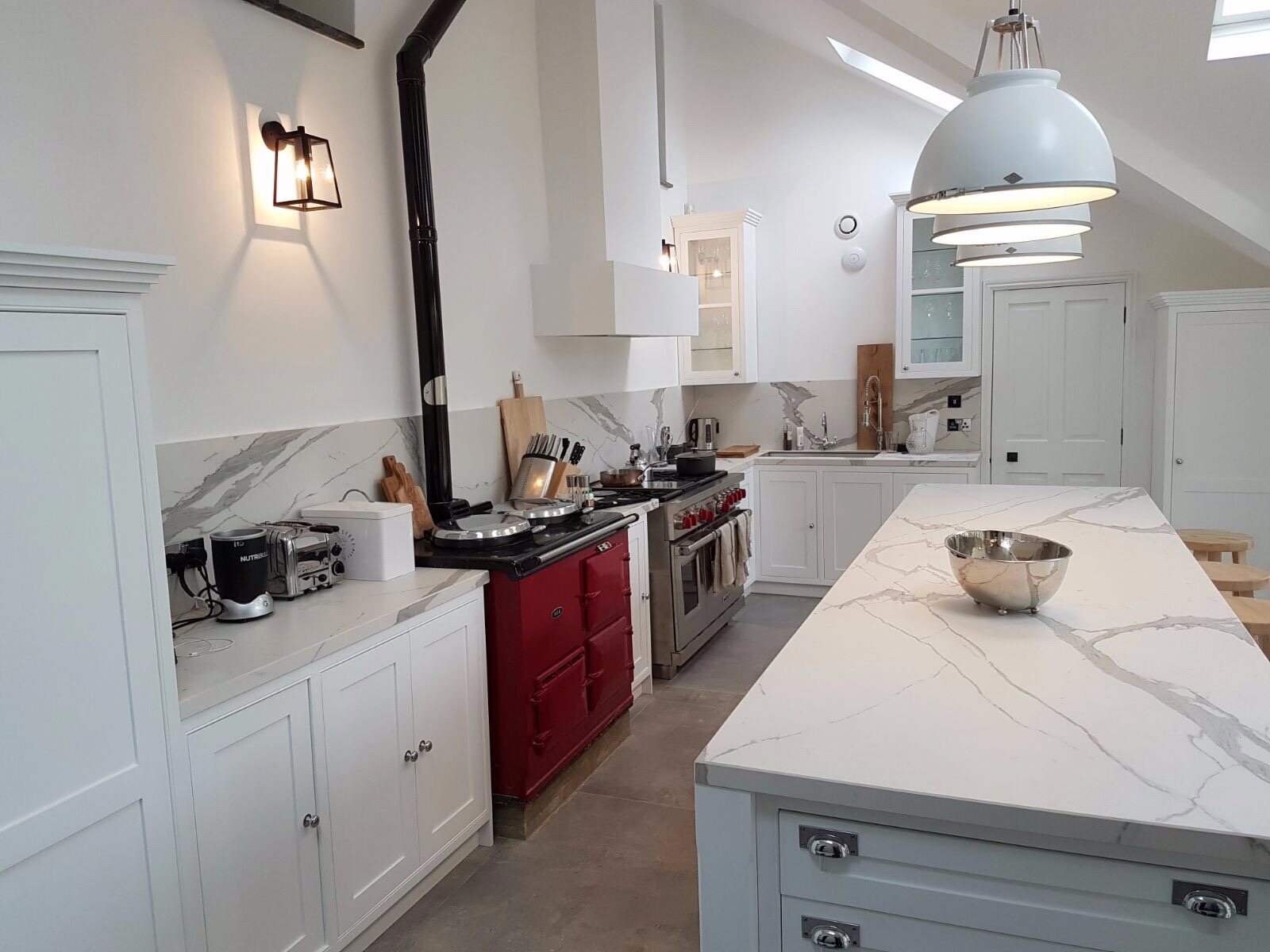 Jasna kuchnia w stylu rustykalnym z czerwonym piecem kaflowym, marmurowym blatem i białymi szafkami, oświetlona wiszącymi lampami.