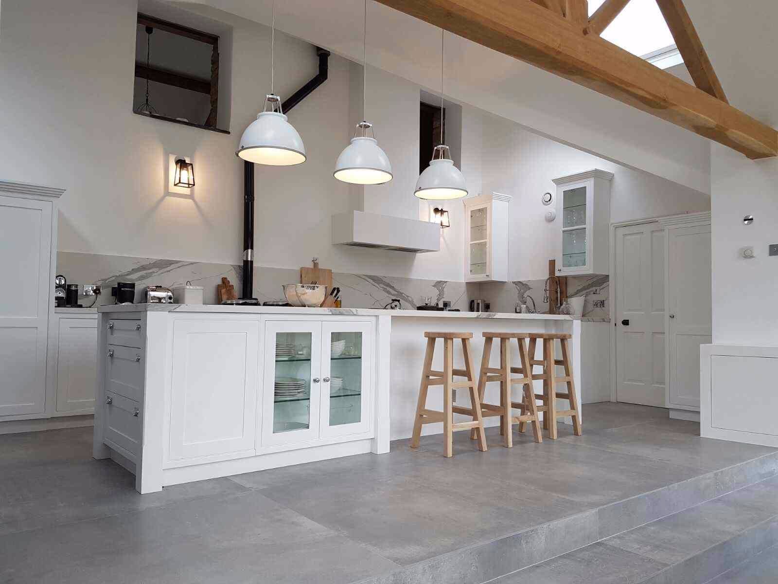 Jasna kuchnia z wyspą, białymi szafkami z przeszklonymi frontami, marmurowym blatem i drewnianymi stołkami barowymi, oświetlona trzema lampami wiszącymi i światłem z okna dachowego.