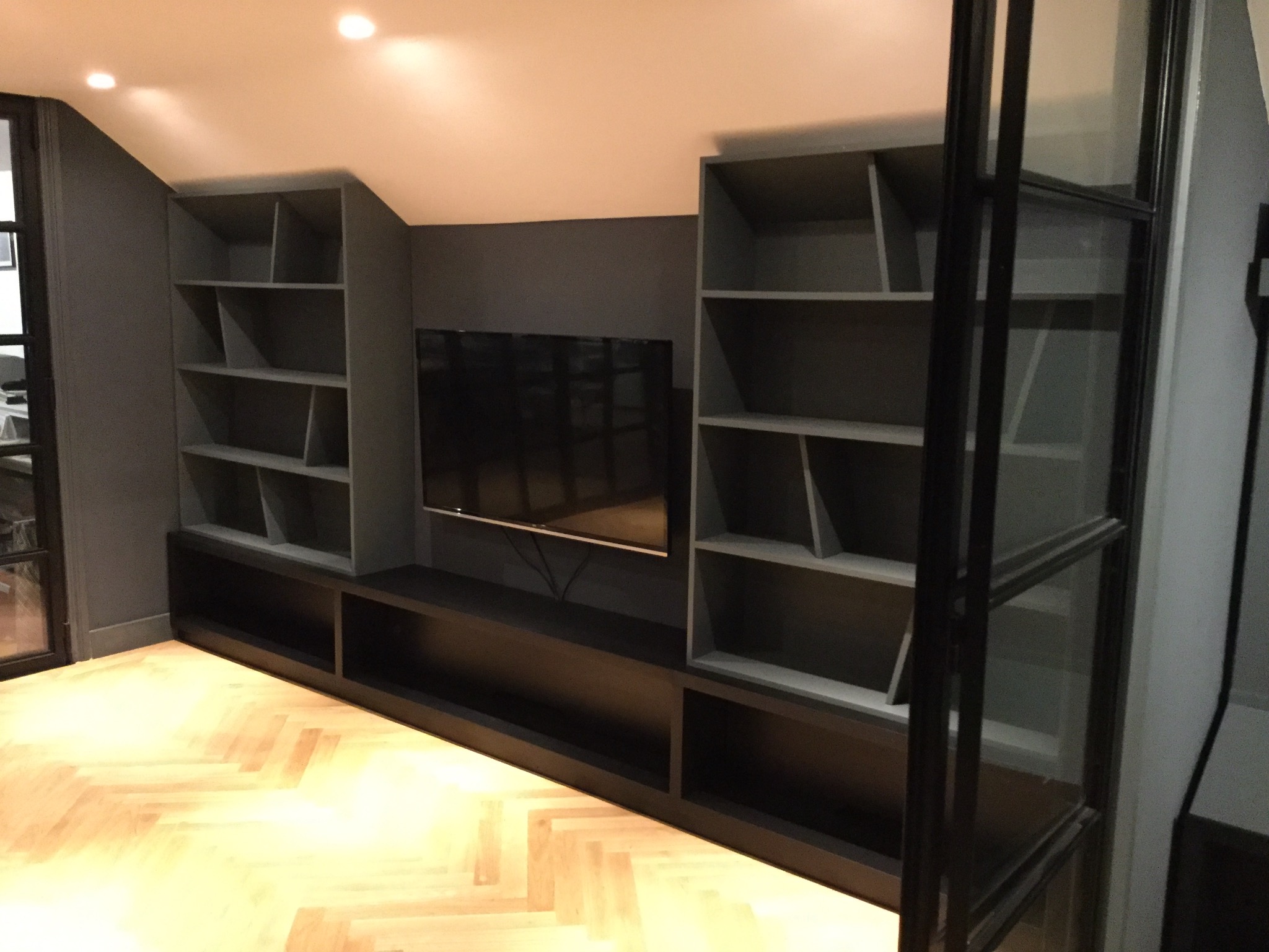 Szara zabudowa meblowa pod skosem z otwartymi półkami i miejscem na telewizor, parkiet w jodełkę na podłodze.