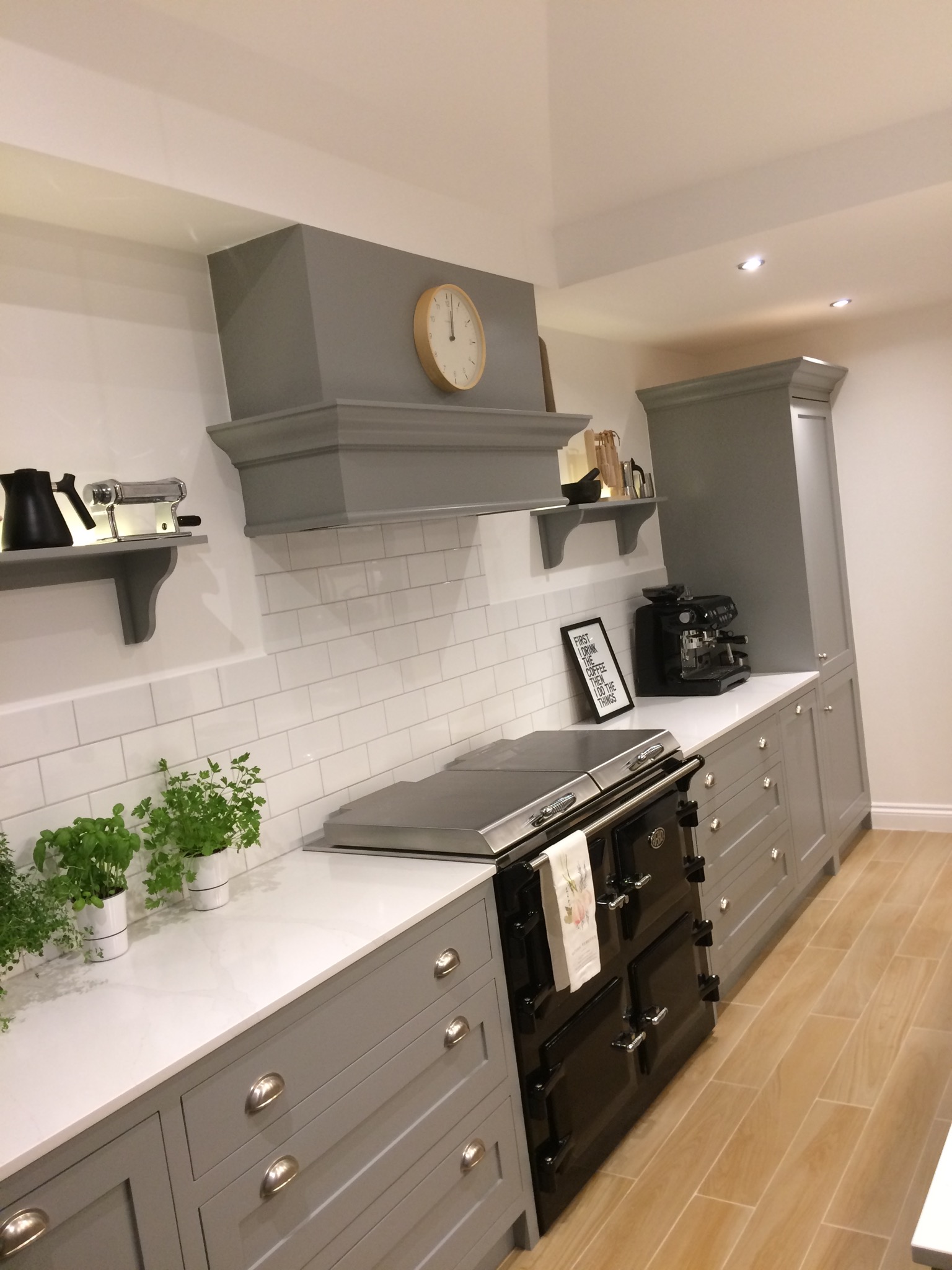 Szara kuchnia na wymiar z czarnym piecem kaflowym, białymi płytkami metra i marmurowym blatem, uzupełniona o zioła w doniczkach i dekoracje na półkach.
