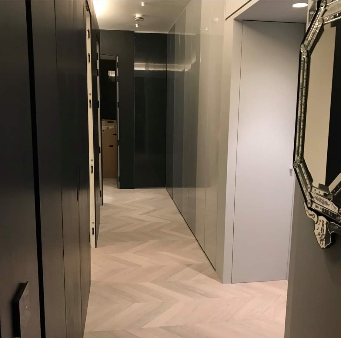 Wąski korytarz z zabudową szaf w kolorach szarości i czerni, parkiet ułożony we wzór jodełki francuskiej, dekoracyjne lustro na ścianie po prawej.