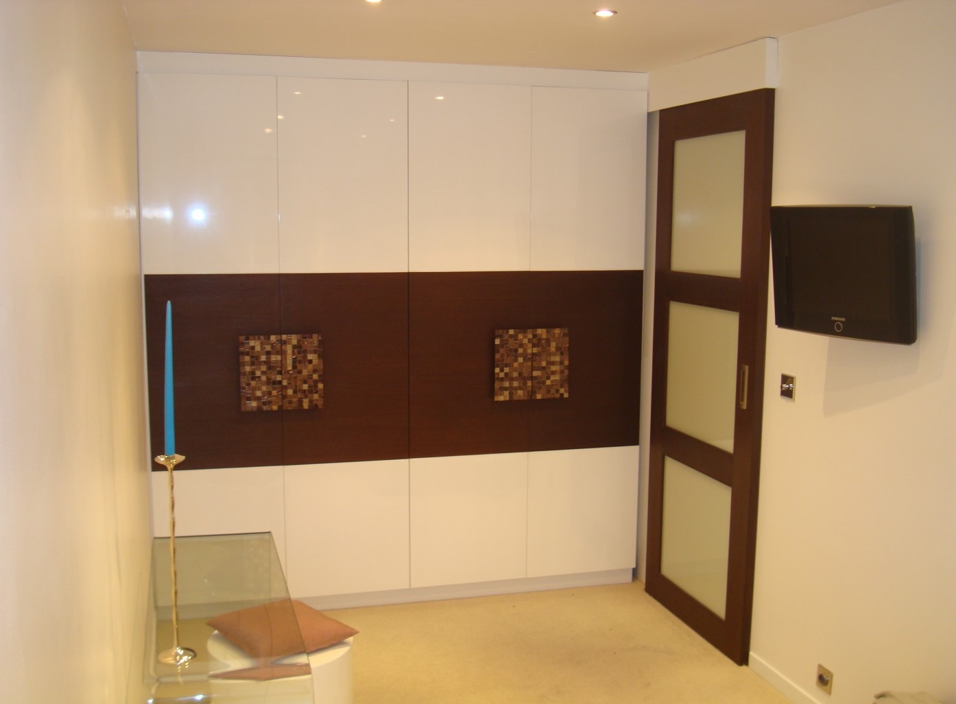 Biało-brązowa szafa wnękowa z dekoracyjnymi, kwadratowymi mozaikami, obok drzwi przesuwnych z mlecznym szkłem i telewizor na ścianie.