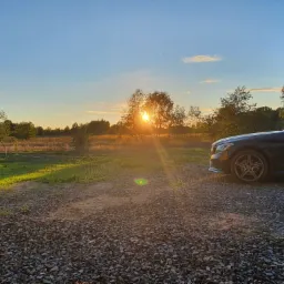 Czarny samochód osobowy zaparkowany na żwirowym podłożu, na tle zachodzącego słońca i drzew.