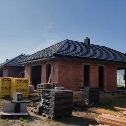 Budowa domów jednorodzinnych w stanie surowym zamkniętym: widoczne mury z bloczków, czarne dachówki na ukończonych dachach, przygotowana więźba dachowa na jednym z budynków oraz palety...