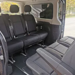 Wnętrze busa z trzema rzędami czarnych foteli, widok z otwartych drzwi bocznych na ulicę.