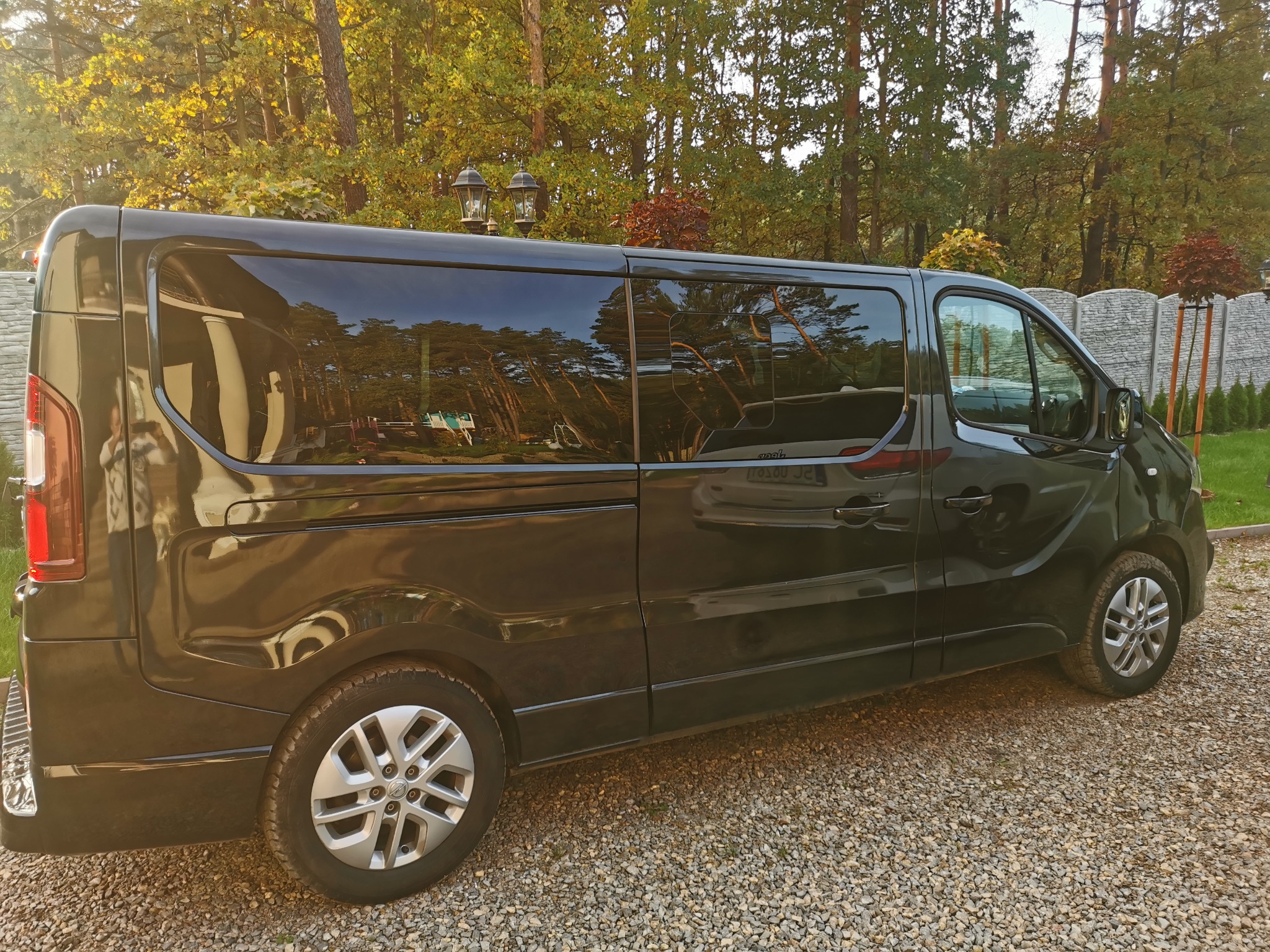 Czarny van, widok z boku, zaparkowany na żwirowym podłożu, z widocznym odbiciem drzew w oknach.