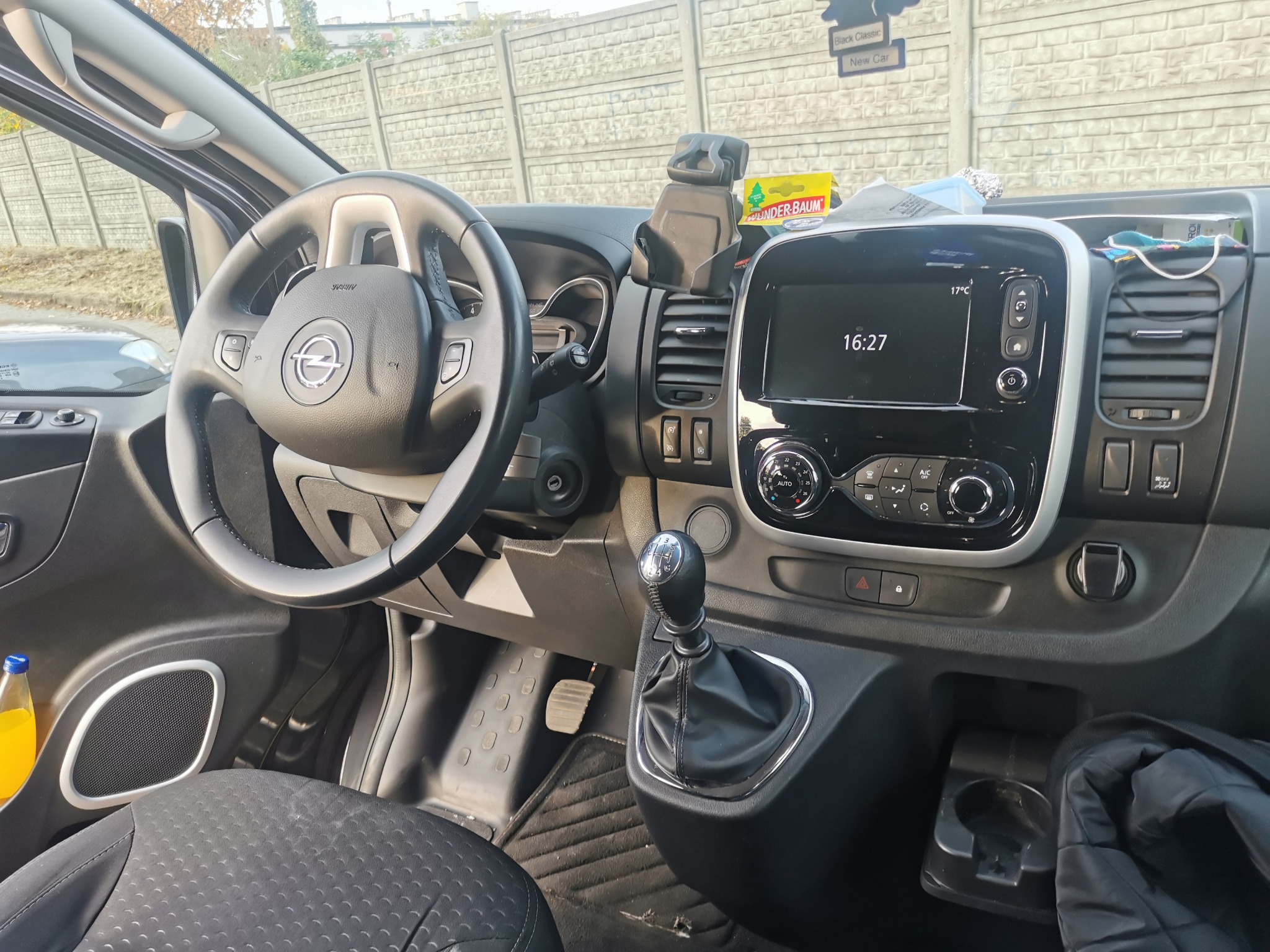 Wnętrze samochodu dostawczego Opel Vivaro z widoczną kierownicą z logo, deską rozdzielczą z ekranem, dźwignią zmiany biegów, oraz butelką z napojem w drzwiach.