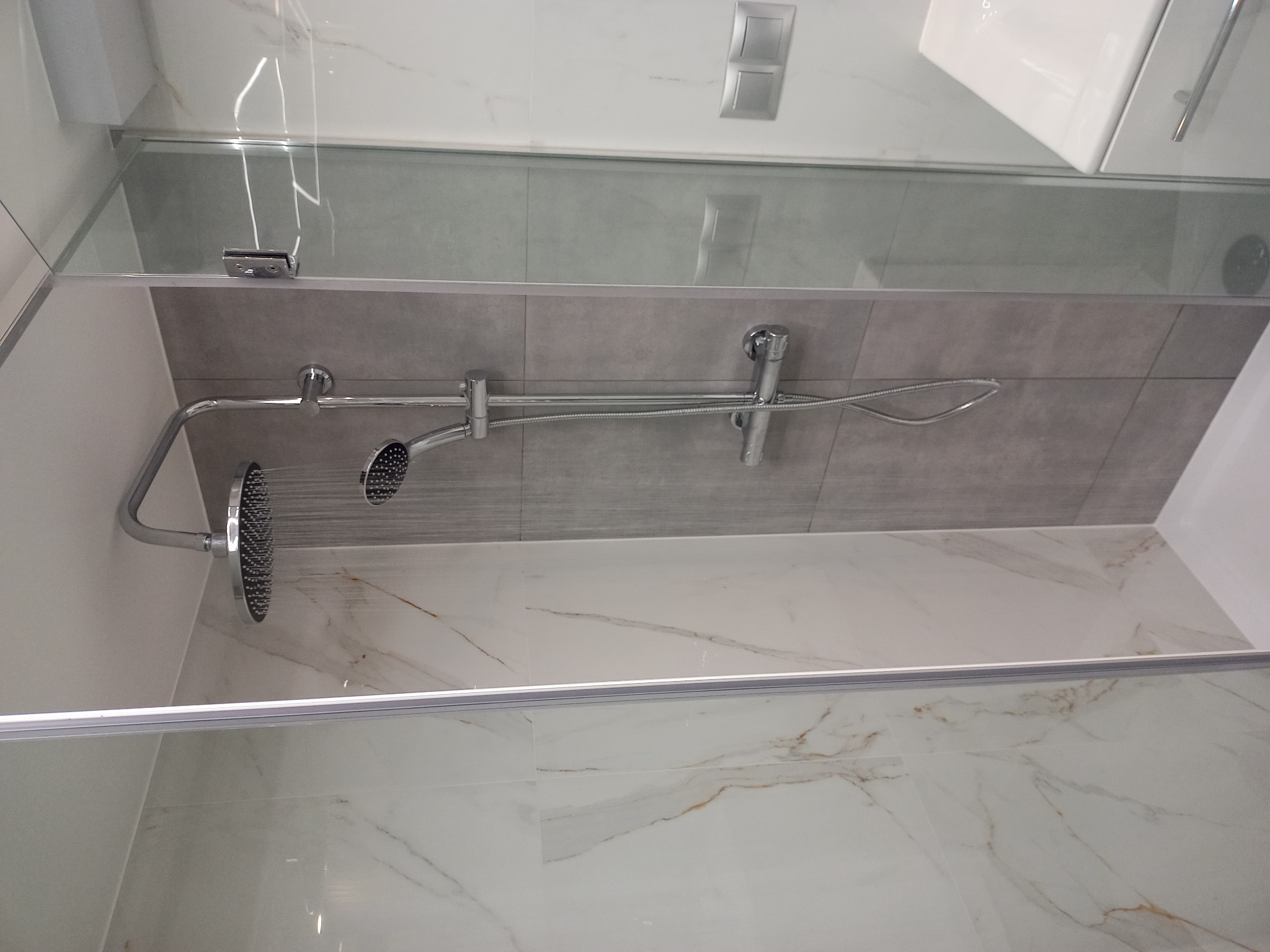 Szklana kabina prysznicowa z deszczownicą i baterią termostatyczną, wykończona płytkami imitującymi marmur i szarymi płytkami na ścianie, widok z góry na wnętrze kabiny.