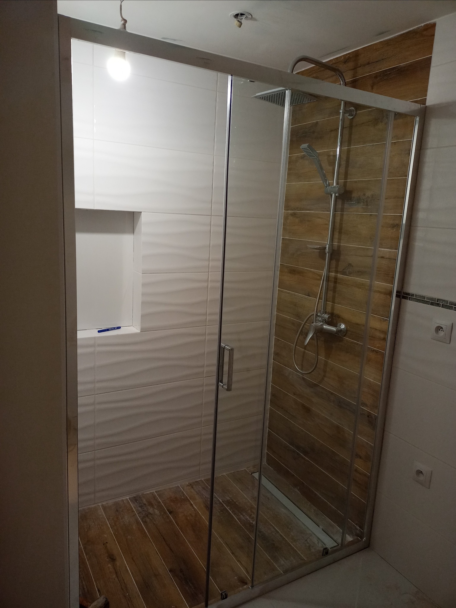 Kabina prysznicowa z deszczownicą i baterią, wykończona płytkami imitującymi drewno i białymi, falistymi płytkami, z drewnianą podłogą i wnęką w ścianie z niebieskim pisakiem.