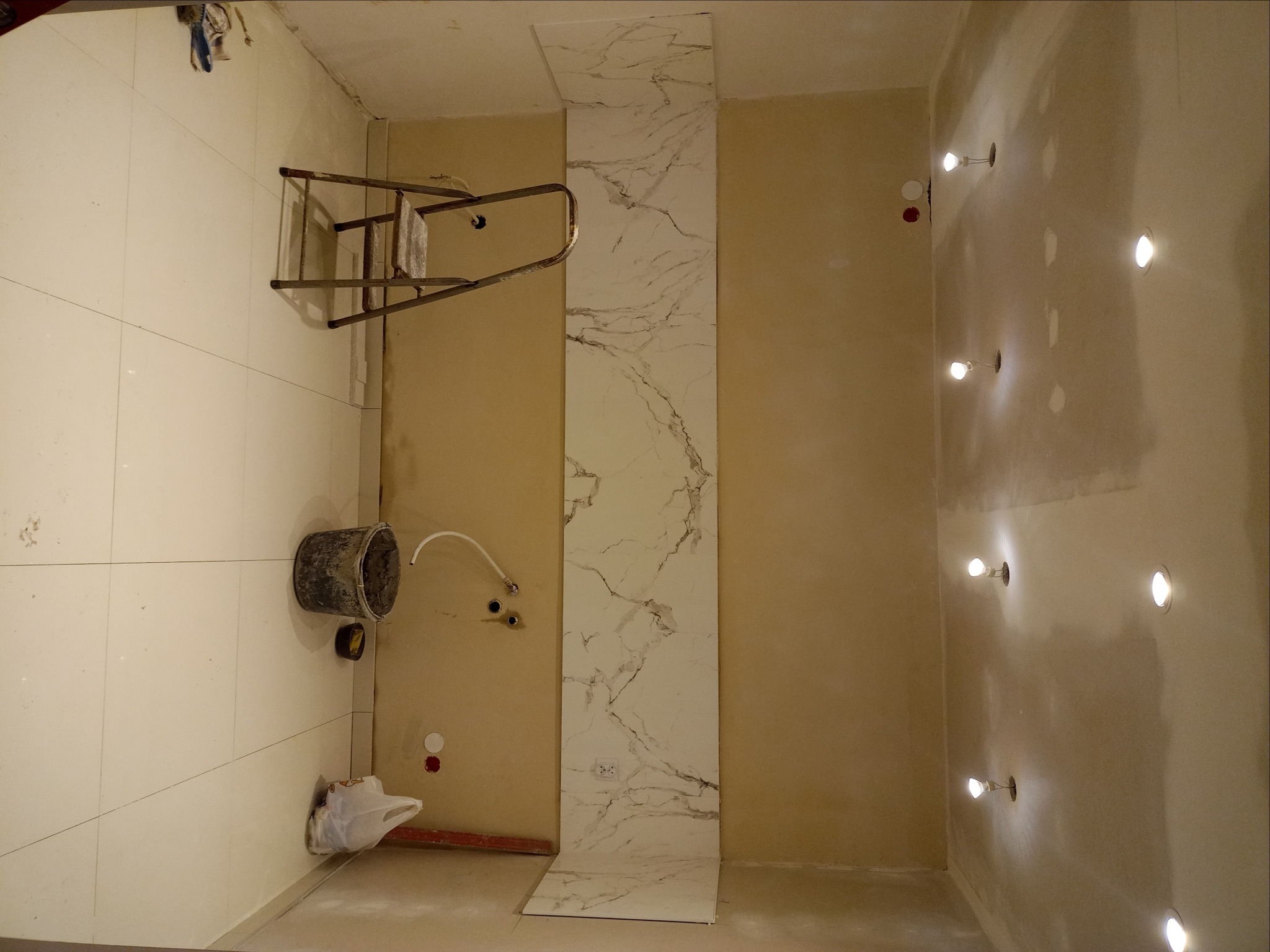 Niedokończona łazienka z położonymi płytkami imitującymi marmur na ścianie, widoczne punkty świetlne w suficie, drabina i wiadro na podłodze.