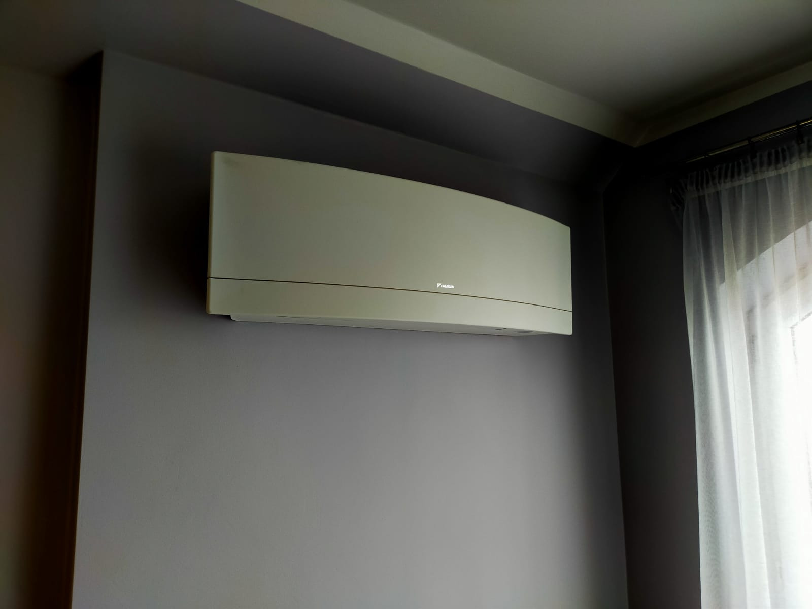 Klimatyzator Daikin zamontowany na szarej ścianie w pokoju z firanką w oknie.