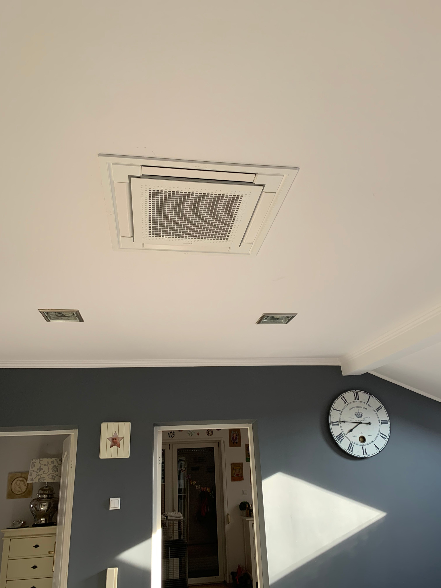 Klimatyzator kasetonowy zamontowany w suficie podwieszanym w pomieszczeniu mieszkalnym, widoczne kratki wentylacyjne i elementy oświetlenia sufitowego.