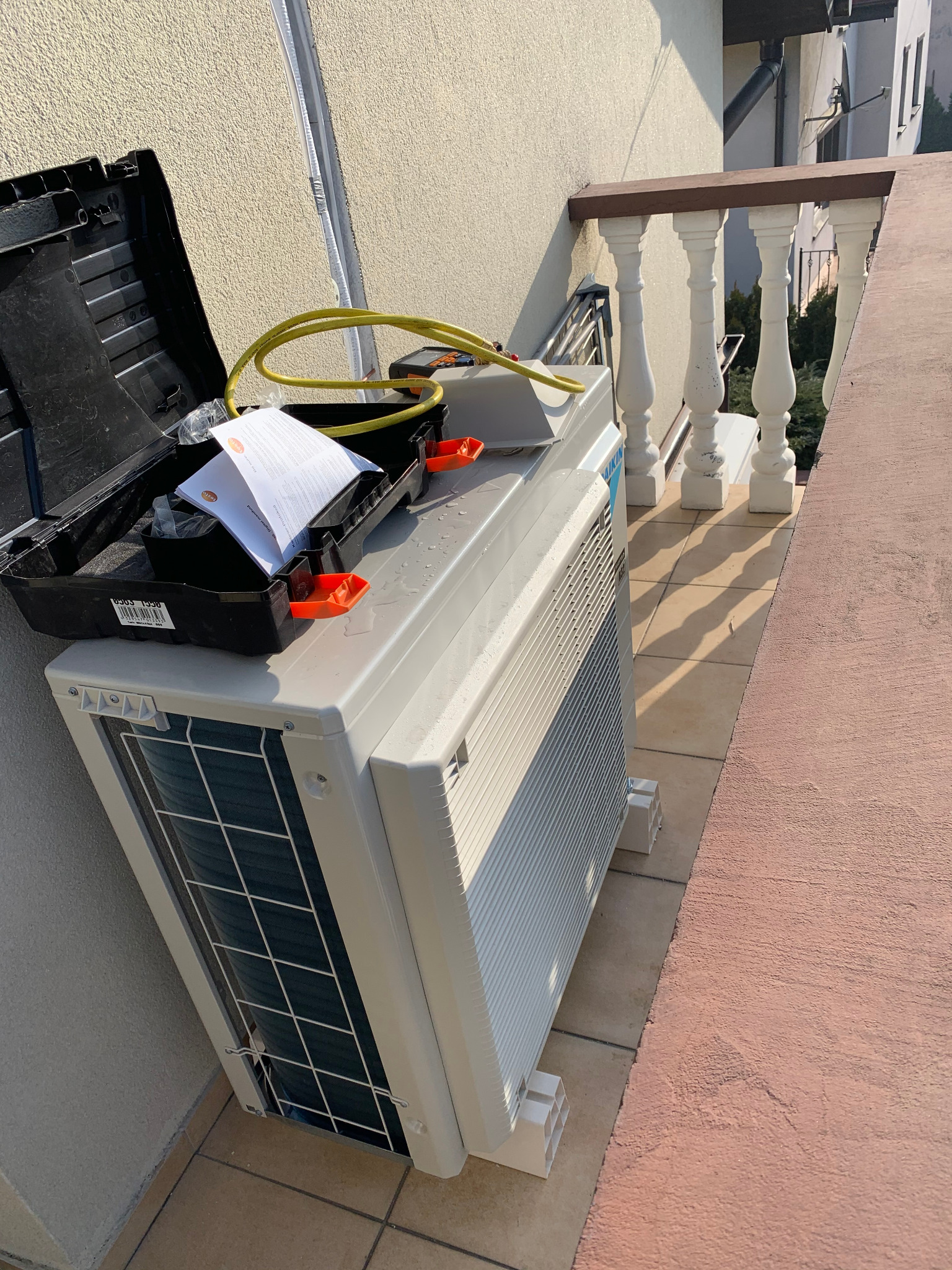 Zewnętrzna jednostka klimatyzatora Daikin na balkonie, obok otwarta skrzynka narzędziowa z przewodami i dokumentacją, widoczne elementy montażowe i przygotowanie do instalacji.