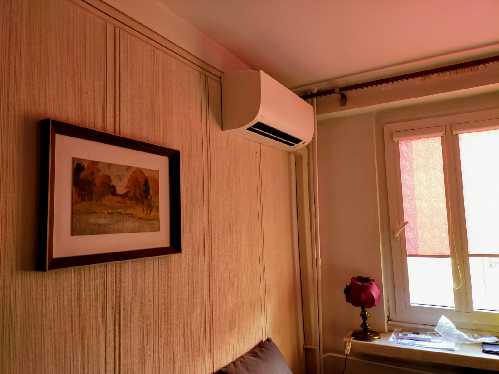Biały klimatyzator ścienny zamontowany w pokoju z tapetą w pionowe paski, obrazem w brązowej ramie i oknem z roletą.