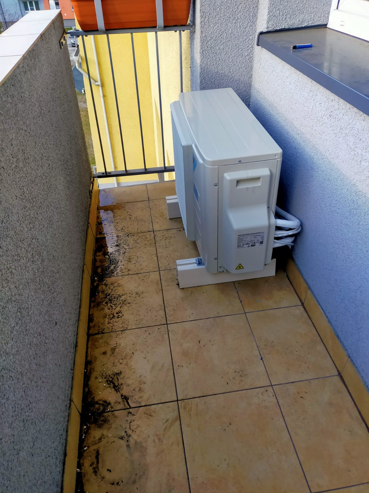 Zewnętrzna jednostka klimatyzacji zamontowana na balkonie z żółtymi płytkami, widoczne przewody i podpory montażowe, ślady wilgoci na podłodze balkonu.