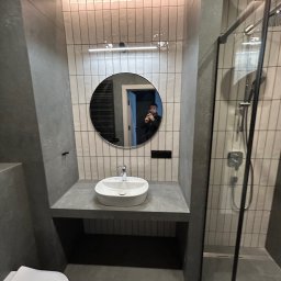 Progress Glazura - Nowoczesna łazienka w szarościach: okrągłe lustro z odbiciem osoby, biała umywalka nablatowa, kabina prysznicowa z czarnymi ramami i pionowe płytki.