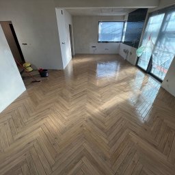 Progress Glazura - Parkiet ułożony w jodełkę w jasnym, przestronnym pokoju. Widoczne przygotowania do dalszych prac wykończeniowych, okna zabezpieczone folią. Podłoga błyszcząca.