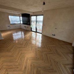 Progress Glazura - Ukończona podłoga z parkietu ułożonego w jodełkę w jasnym, przestronnym pokoju. Ściany przygotowane do malowania, okna zabezpieczone folią malarską.