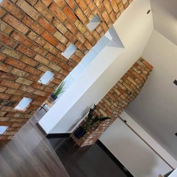 Progress Glazura - Ceglana ściana z wnękami, drewniana podłoga, czarne płytki, roślina doniczkowa, niebieska folia na podłodze, widok z dołu.