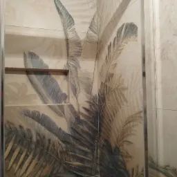 Beżowe płytki z motywem liści tropikalnych na ścianie, z wnęką na kosmetyki i metalową listwą narożną.