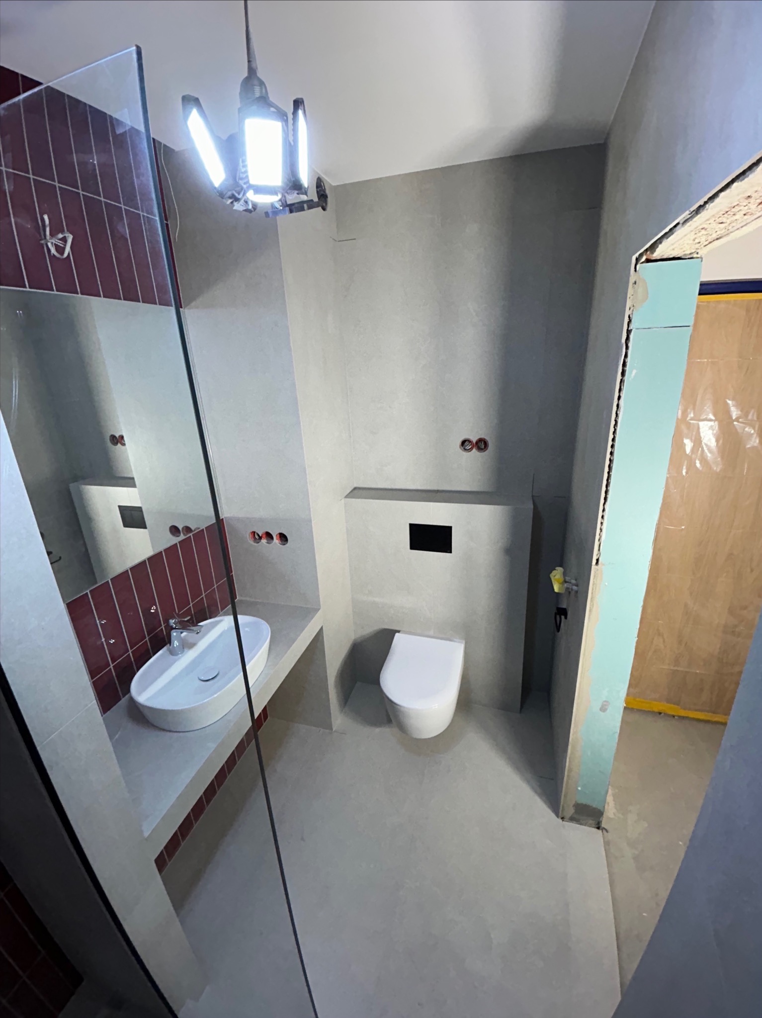 Wnętrze łazienki w trakcie prac wykończeniowych: widoczna umywalka, toaleta, lustro i płytki w odcieniach szarości i bordo, oświetlenie robocze, surowy stan wykończenia.