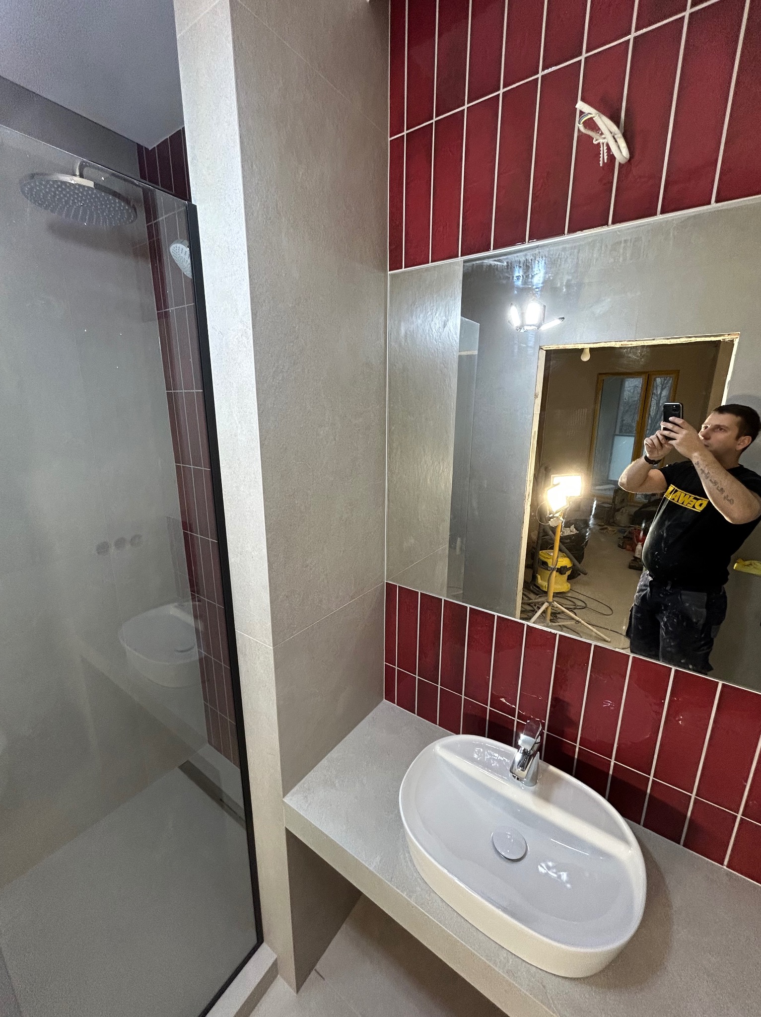 Nowoczesna łazienka z kabiną prysznicową typu walk-in, umywalką nablatową i czerwoną glazurą. W lustrze odbija się osoba robiąca zdjęcie.