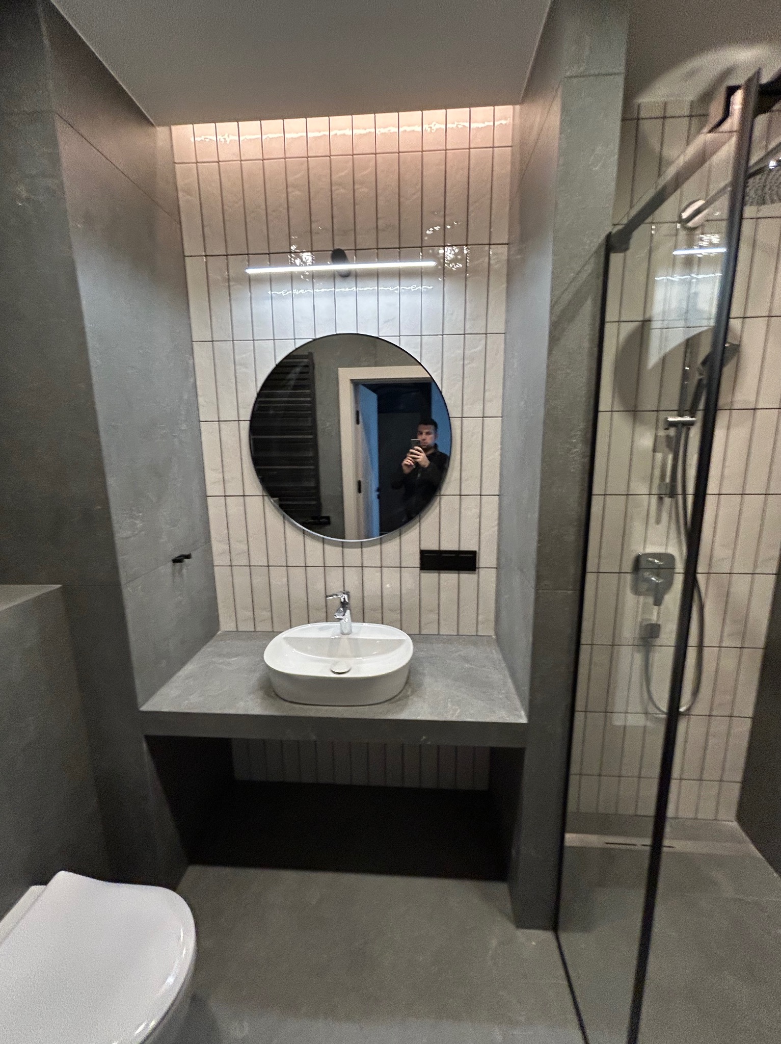 Nowoczesna łazienka w szarościach: okrągłe lustro z odbiciem osoby, biała umywalka nablatowa, kabina prysznicowa z czarnymi ramami i pionowe płytki.