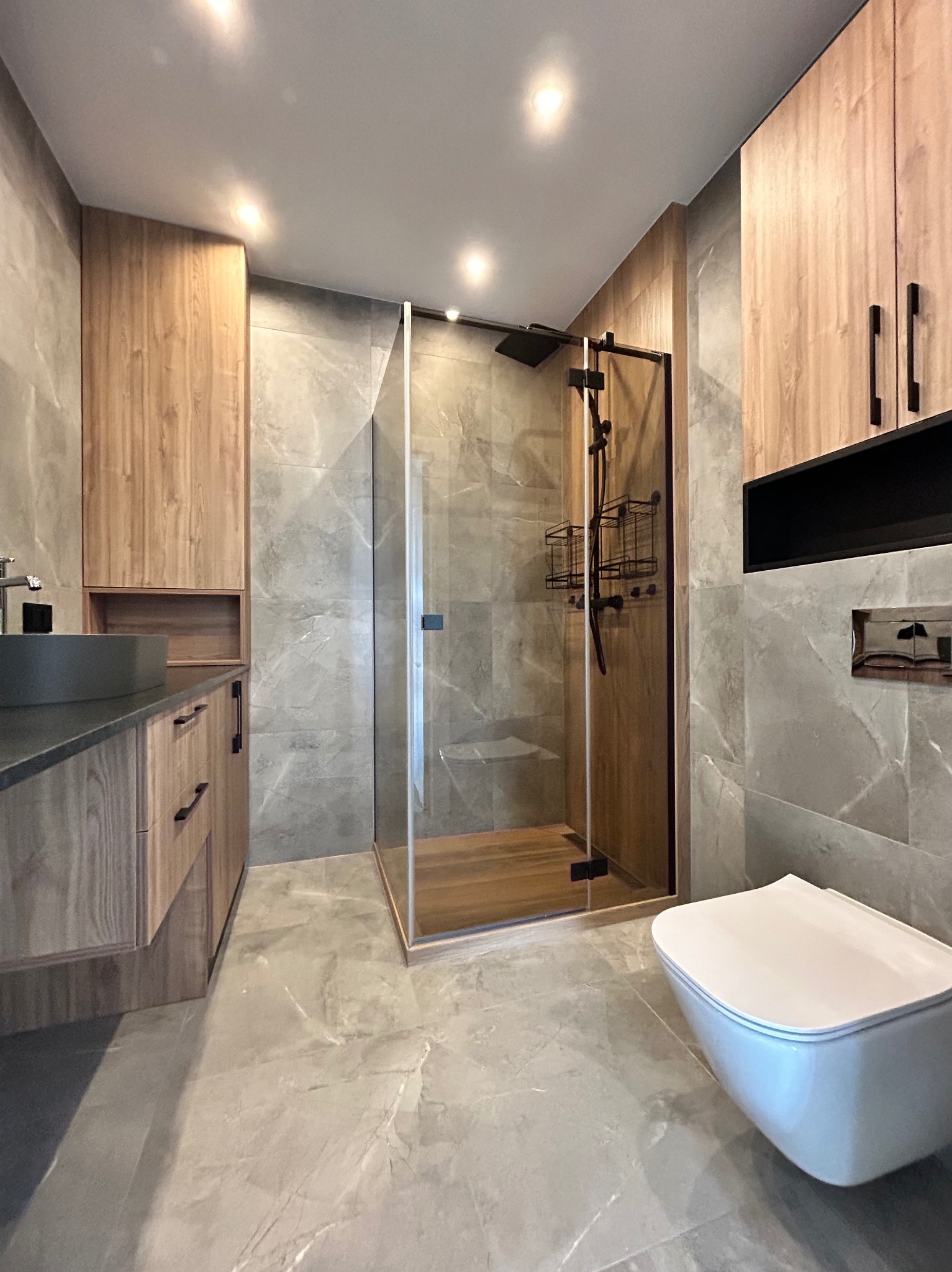 Nowoczesna łazienka z kabiną prysznicową typu walk-in, szarą glazurą i drewnianymi szafkami. Umywalka w kolorze grafitowym. Minimalistyczny design.