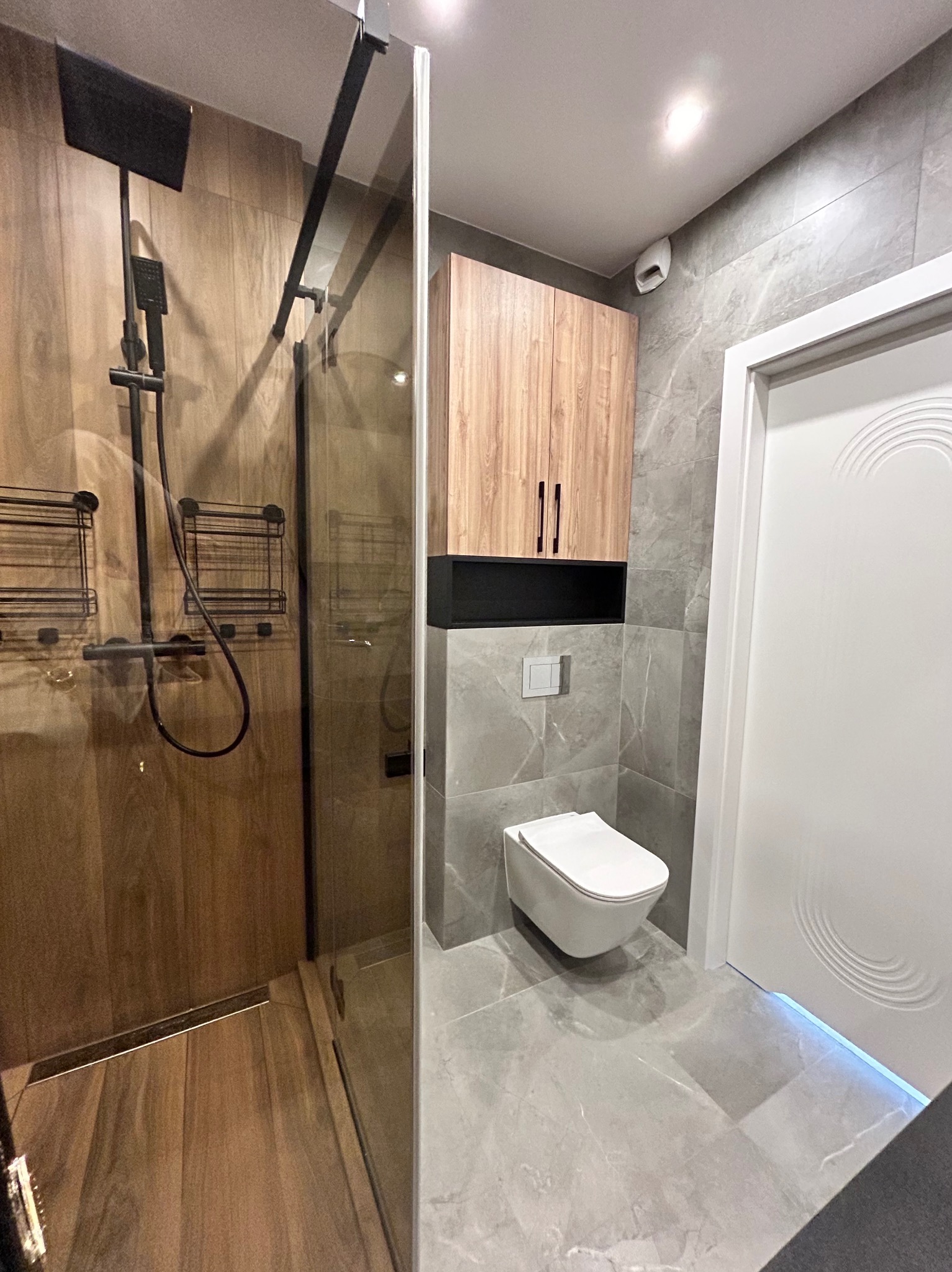 Nowoczesna łazienka z kabiną typu walk-in, drewnopodobnymi płytkami w strefie prysznica i szarymi płytkami na ścianach i podłodze. Podwieszana toaleta i szafka nad nią.