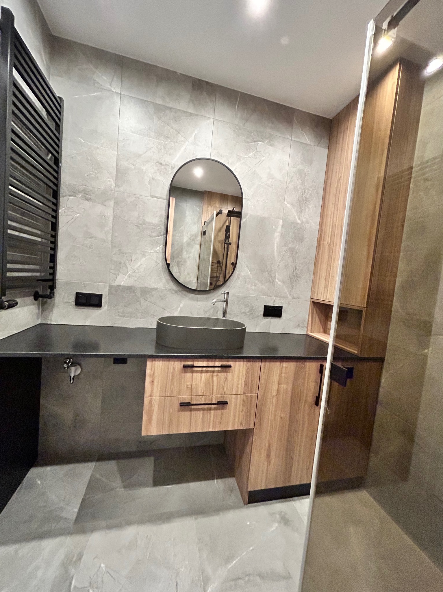 Nowoczesna łazienka z owalnym lustrem, szarą umywalką i drewnianymi szafkami. Widoczny grzejnik drabinkowy i kabina prysznicowa ze szklaną ścianką.