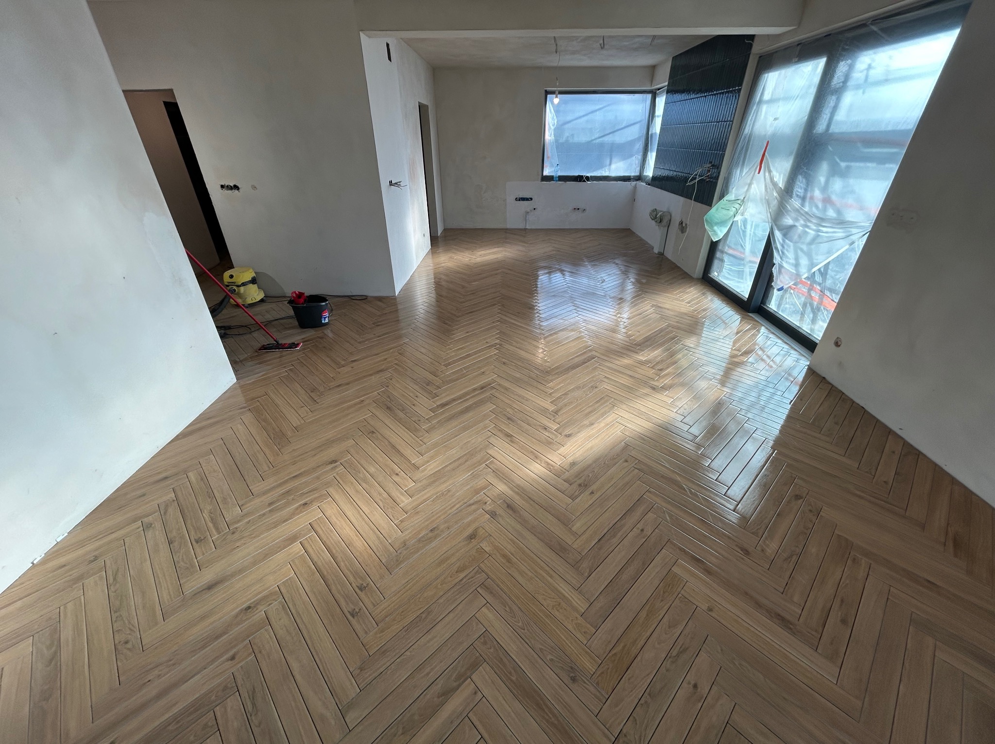 Parkiet ułożony w jodełkę w jasnym, przestronnym pokoju. Widoczne przygotowania do dalszych prac wykończeniowych, okna zabezpieczone folią. Podłoga błyszcząca.