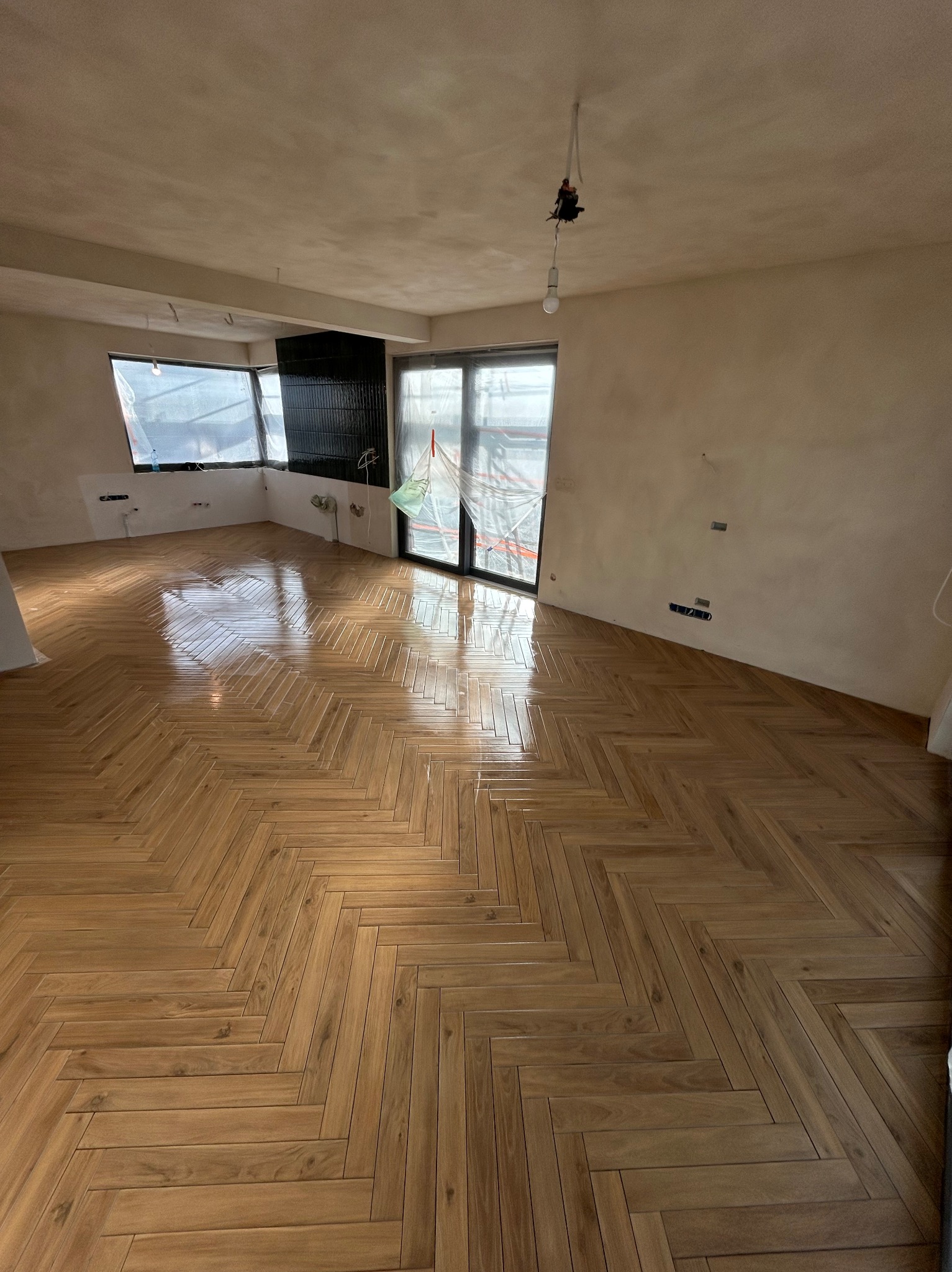 Ukończona podłoga z parkietu ułożonego w jodełkę w jasnym, przestronnym pokoju. Ściany przygotowane do malowania, okna zabezpieczone folią malarską.