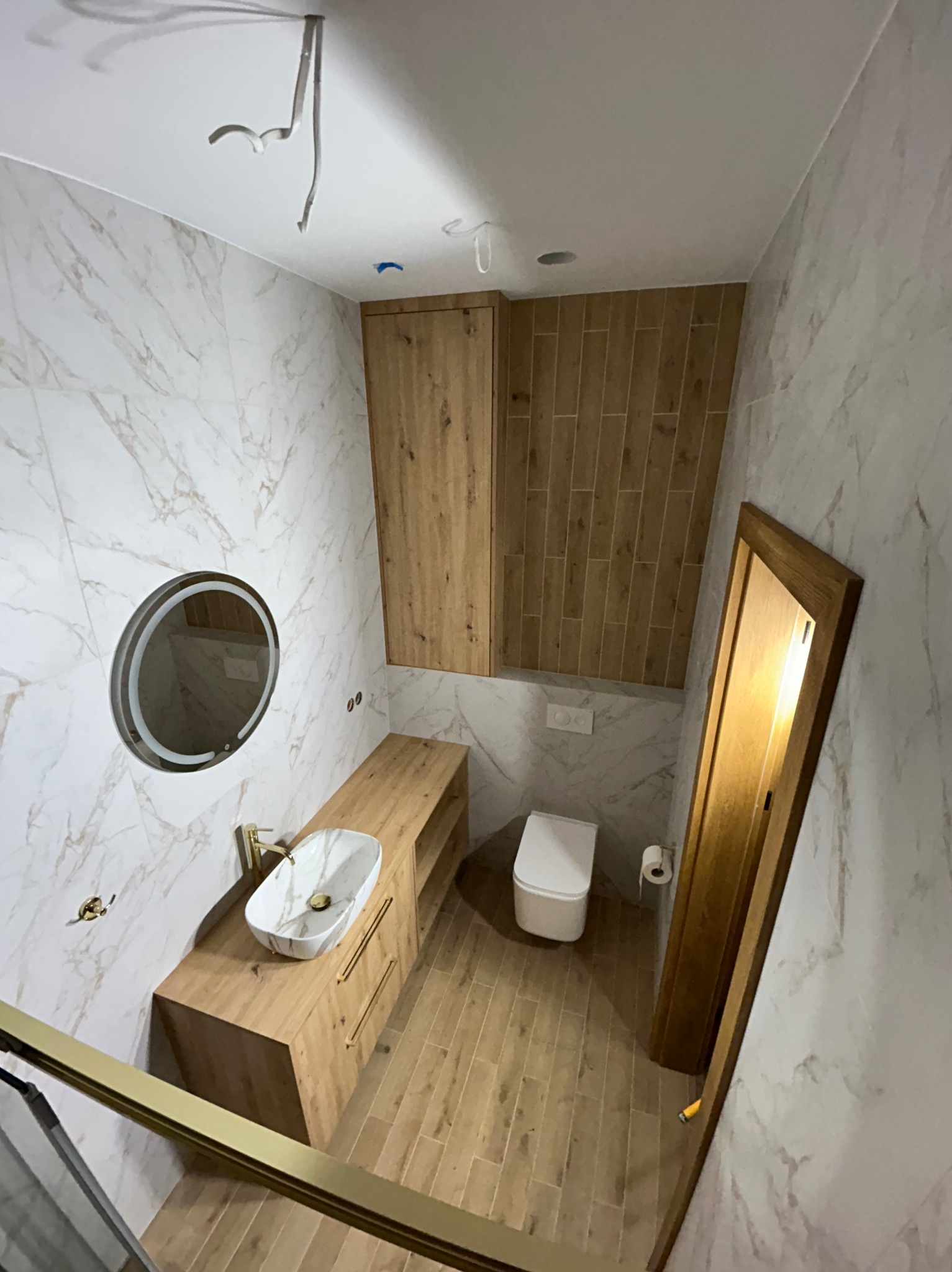 Nowoczesna łazienka po remoncie: marmurowe płytki, drewniane szafki, okrągłe lustro z podświetleniem LED i wisząca toaleta. Ujęcie z góry, widoczne niedokończone oświetlenie.