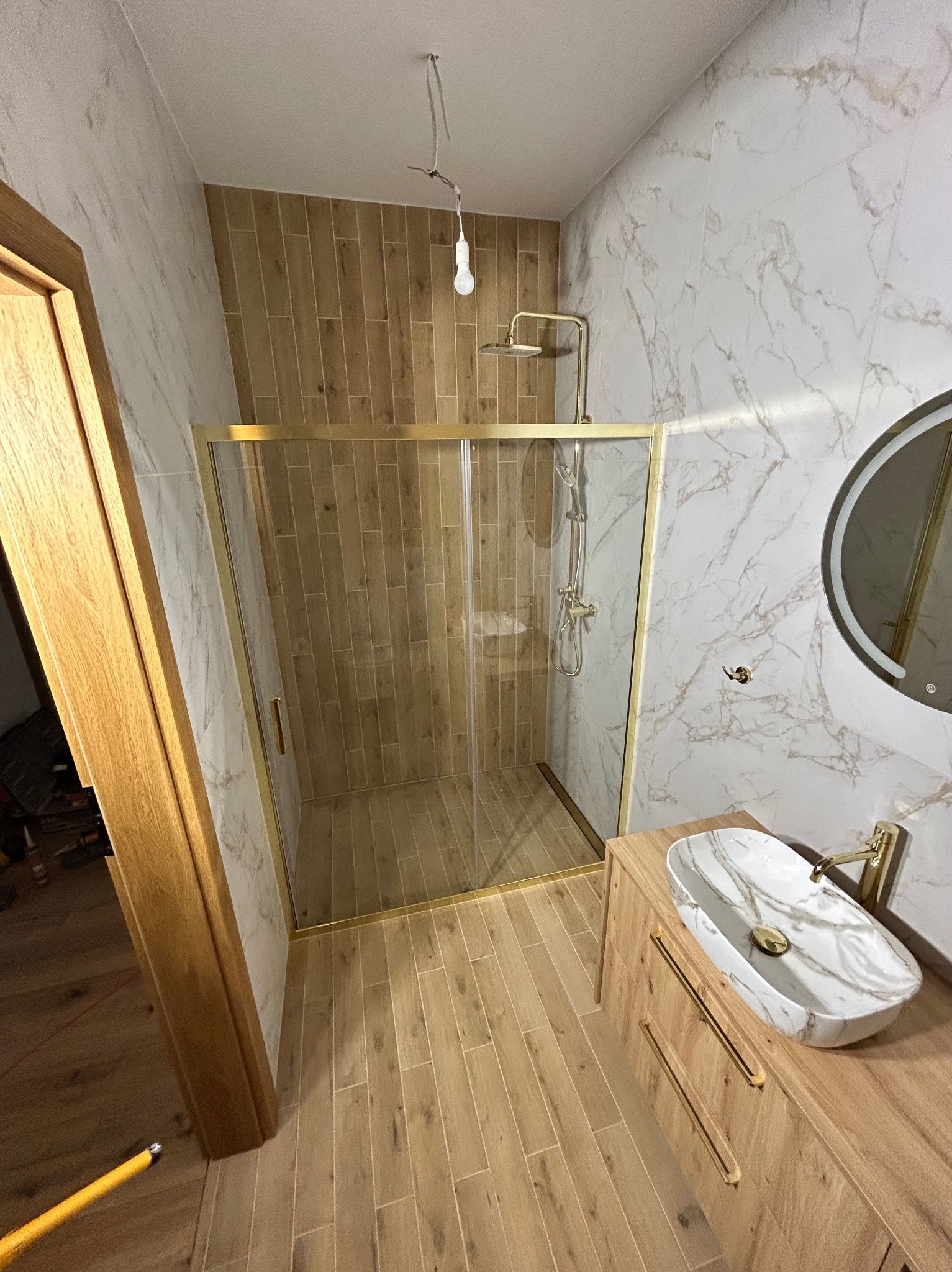 Wykończona łazienka w Wieliczce z kabiną prysznicową w złotej ramie, marmurową umywalką i drewnopodobnymi płytkami. Nowoczesny design.