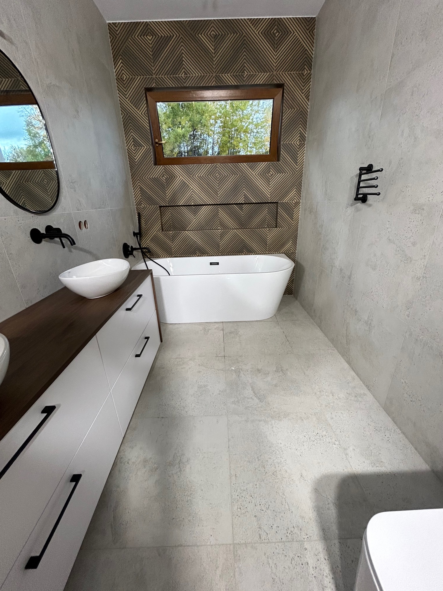 Nowoczesna łazienka z wolnostojącą wanną, umywalką nablatową i geometryczną mozaiką na ścianie. Okno z widokiem na zieleń. Armatura w kolorze czarnym.