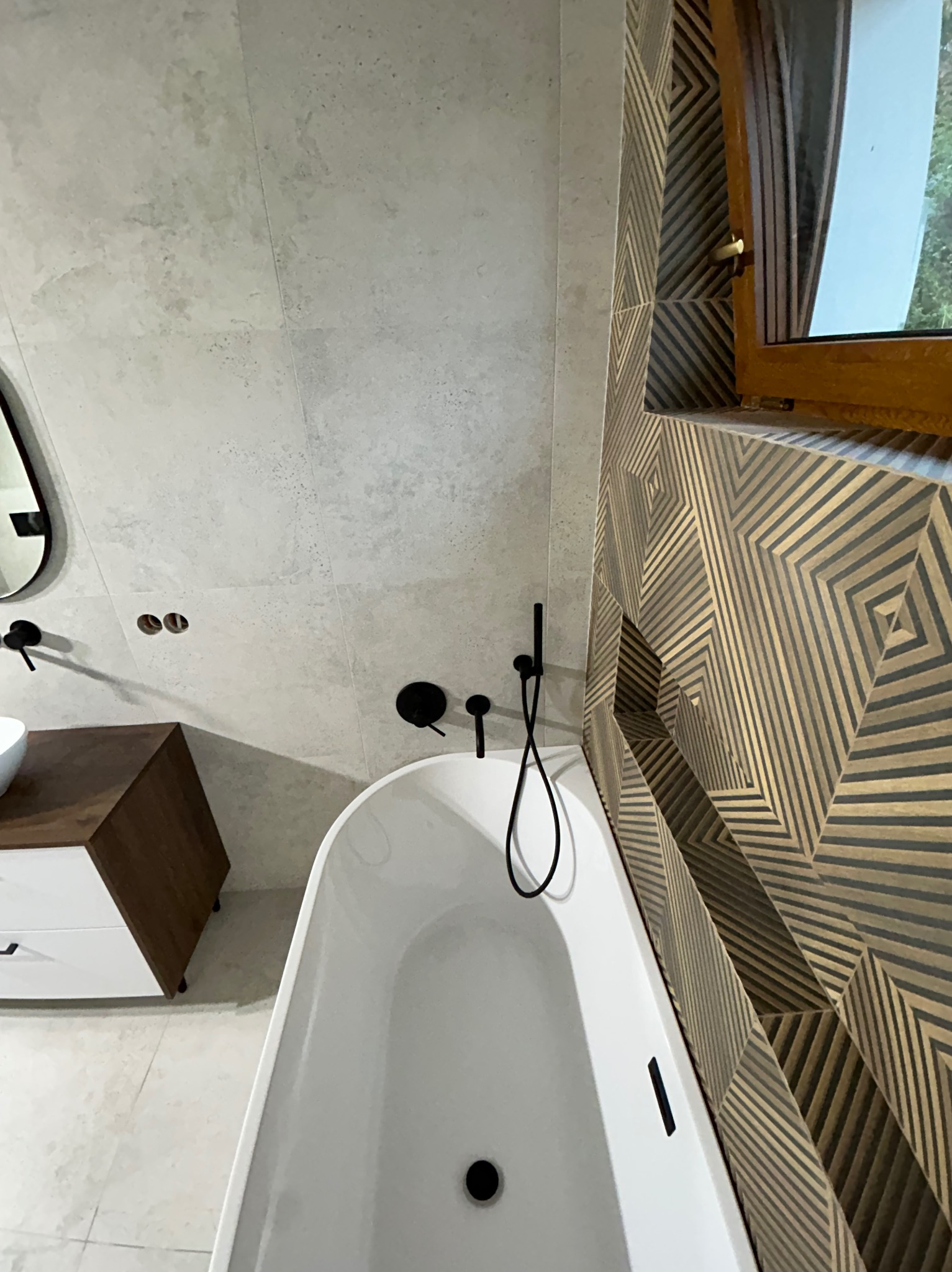 Nowoczesna łazienka z wolnostojącą wanną, czarną armaturą i geometryczną mozaiką na ścianie przy oknie. Minimalistyczny design, jasne płytki i drewniane akcenty.