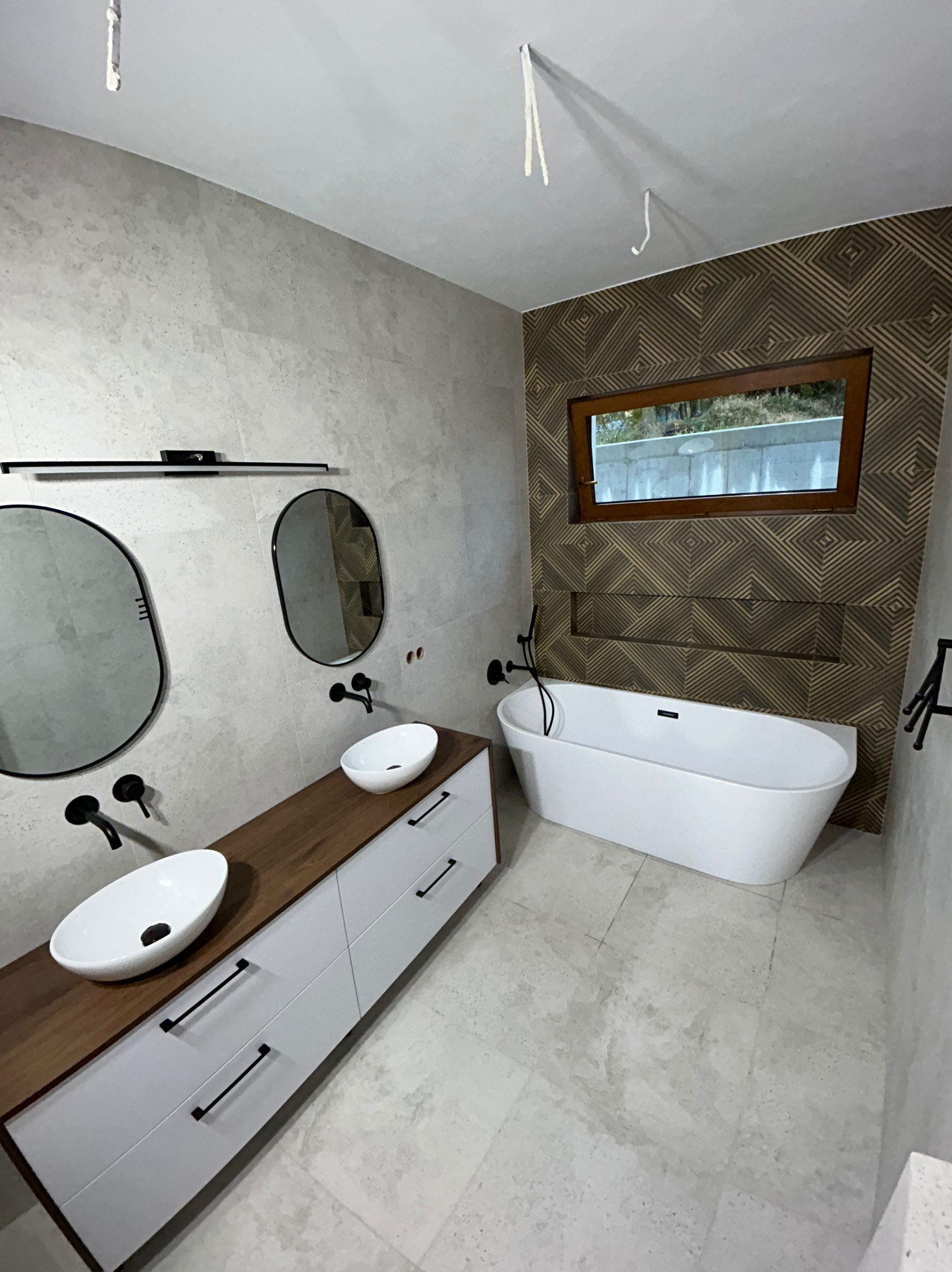 Nowoczesna łazienka z dwoma umywalkami na drewnianym blacie, białą wanną wolnostojącą i geometryczną tapetą. Minimalistyczne czarne baterie i owalne lustra.