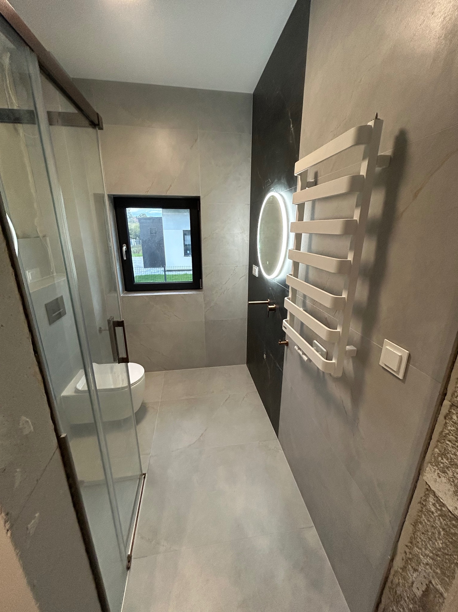 Nowoczesna łazienka z szarymi płytkami, podświetlanym lustrem i grzejnikiem drabinkowym. Kabina prysznicowa ze szklanymi drzwiami, okno z widokiem na zewnątrz.