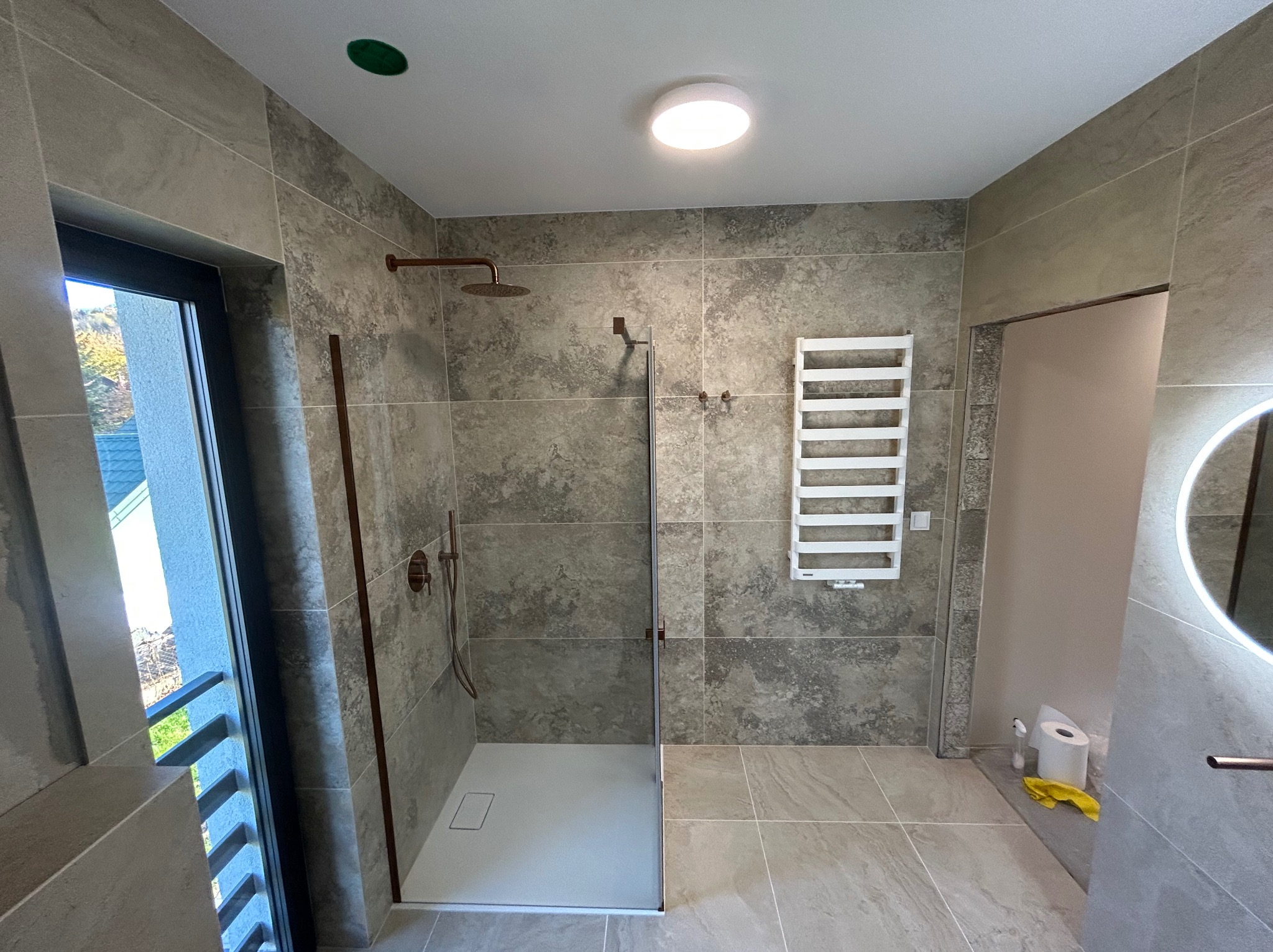 Nowoczesna łazienka z kabiną prysznicową typu walk-in, wykończona płytkami w odcieniach szarości. Widoczny grzejnik drabinkowy i okrągłe lustro z podświetleniem LED.