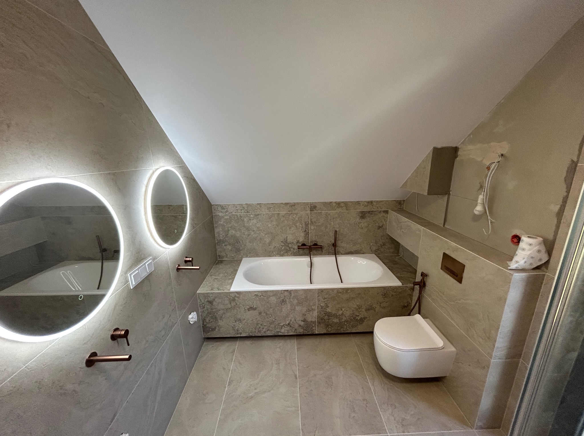 Nowoczesna łazienka na poddaszu z wanną, toaletą i okrągłymi lustrami z podświetleniem LED. Beżowe płytki imitujące kamień. Miedziana armatura.