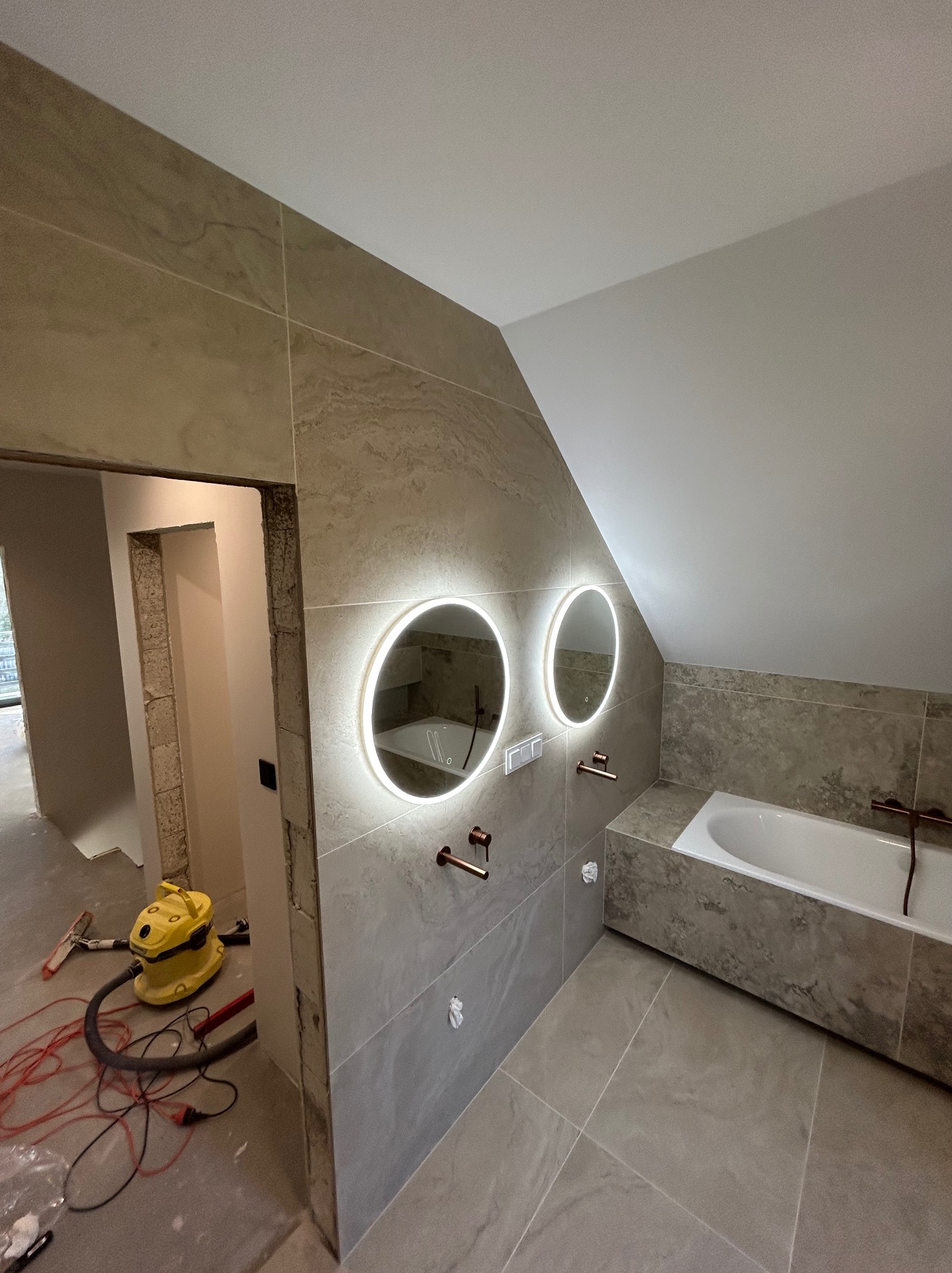 Wykończona łazienka na poddaszu z wanną, okrągłymi lustrami z podświetleniem LED i beżowymi płytkami. Widoczny odkurzacz i narzędzia w przejściu do innego pomieszczenia.