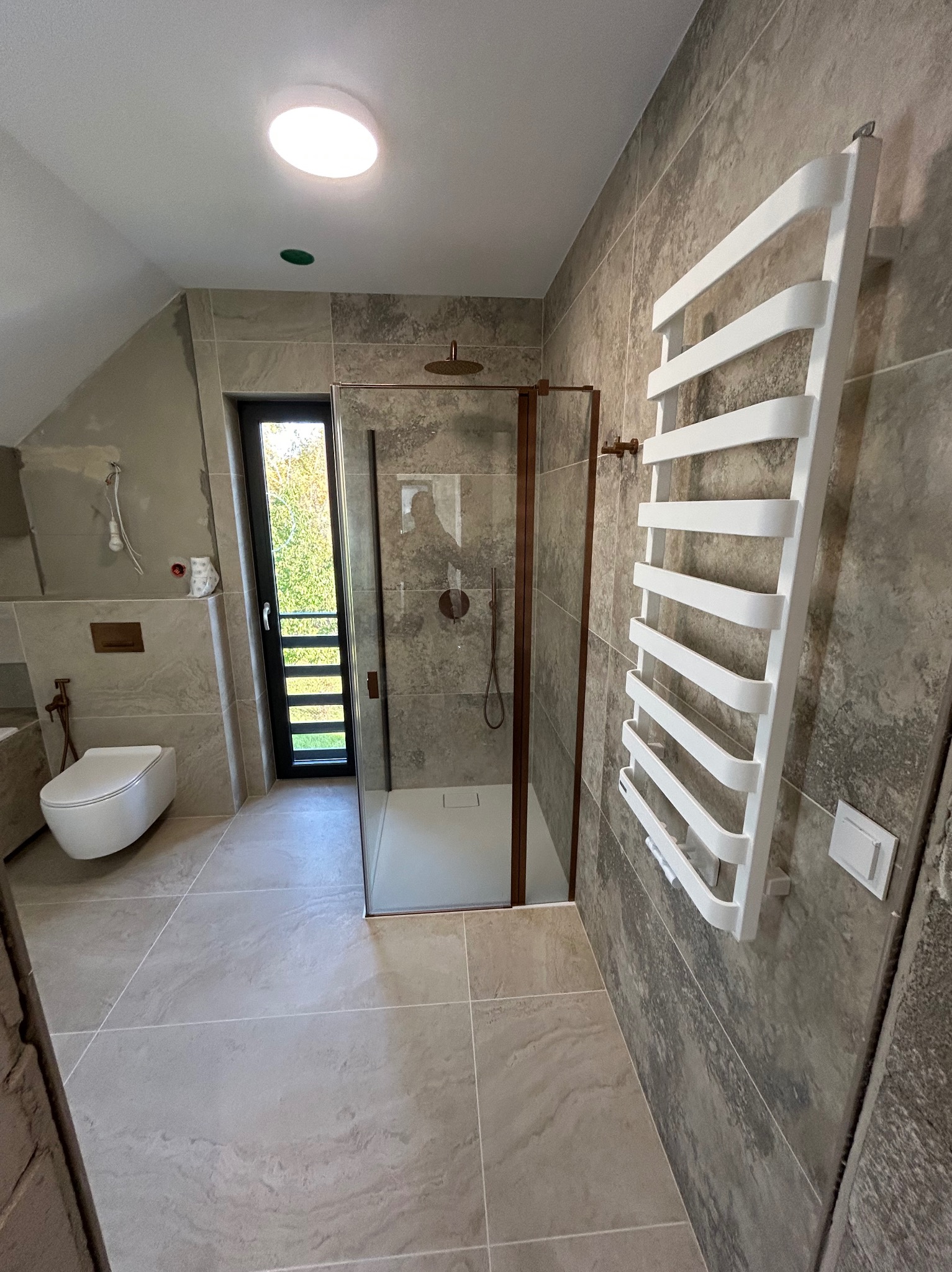 Wykończona łazienka w szarościach z kabiną prysznicową w brązowej ramie, grzejnikiem drabinkowym i podwieszaną toaletą. Część ściany jeszcze nie wykończona.