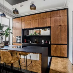 Nowoczesna kuchnia z drewnianymi szafkami, czarnym blatem i złotymi akcentami, oświetlona trzema wiszącymi lampami i doświetlona przez duże okno z widocznymi roślinami doniczkowymi.