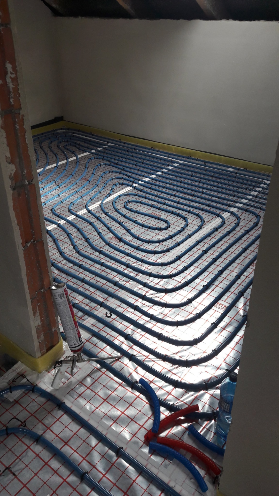 Niebieskie rury grzewcze układane spiralnie na folii z czerwoną siatką w szarej przestrzeni z surową cegłą, widoczna puszka pianki montażowej i fragmenty rur.