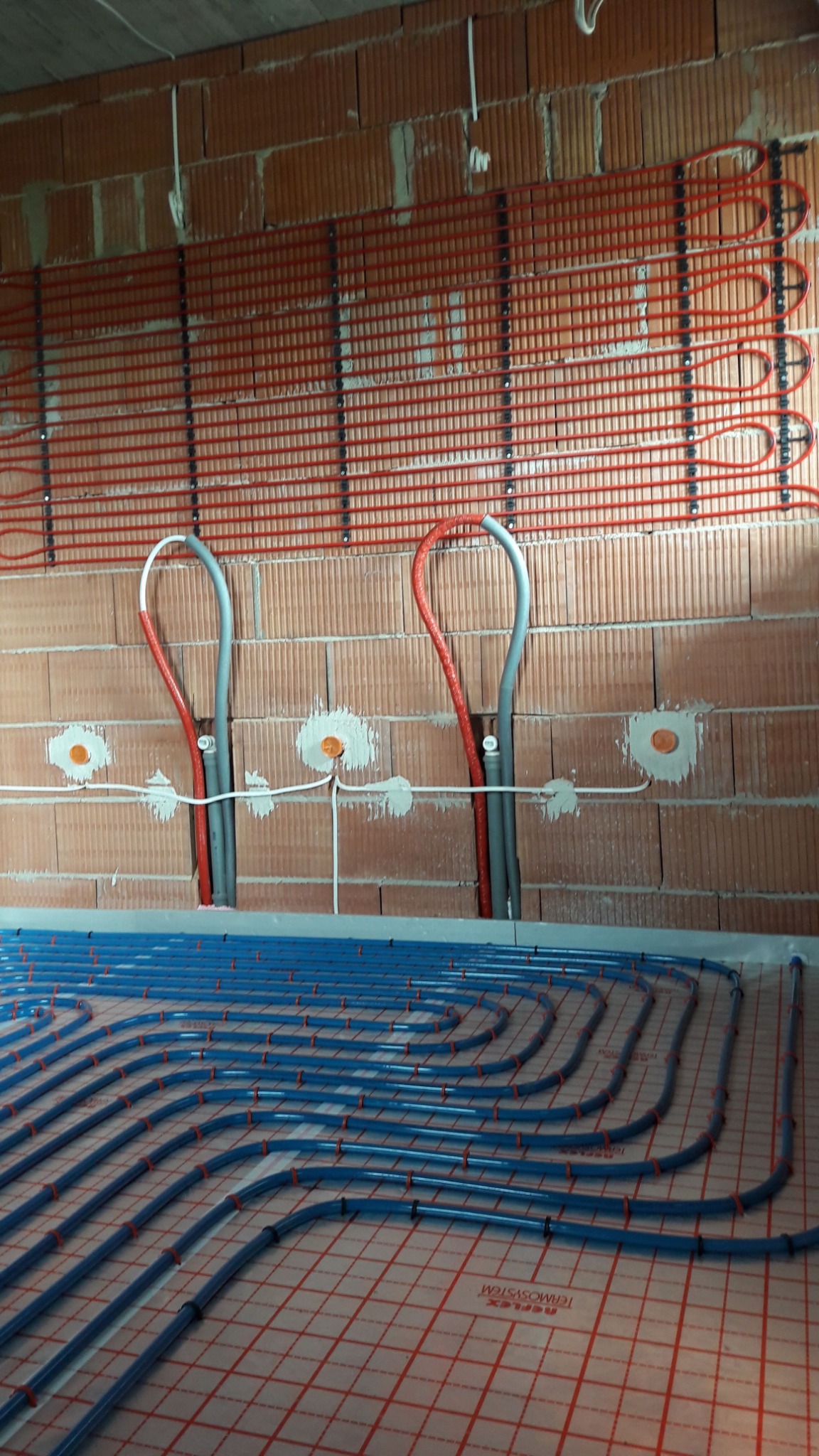 Montaż ogrzewania podłogowego i ściennego w surowym stanie, widoczne rury w kolorze niebieskim i czerwonym, przymocowane do ściany z cegły i podłoża z siatką.