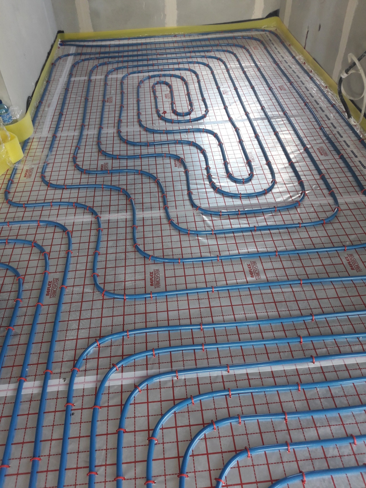 Niebieskie rury ogrzewania podłogowego ułożone na folii z siatką w pomieszczeniu w trakcie instalacji, zabezpieczone żółtą folią przy ścianach.