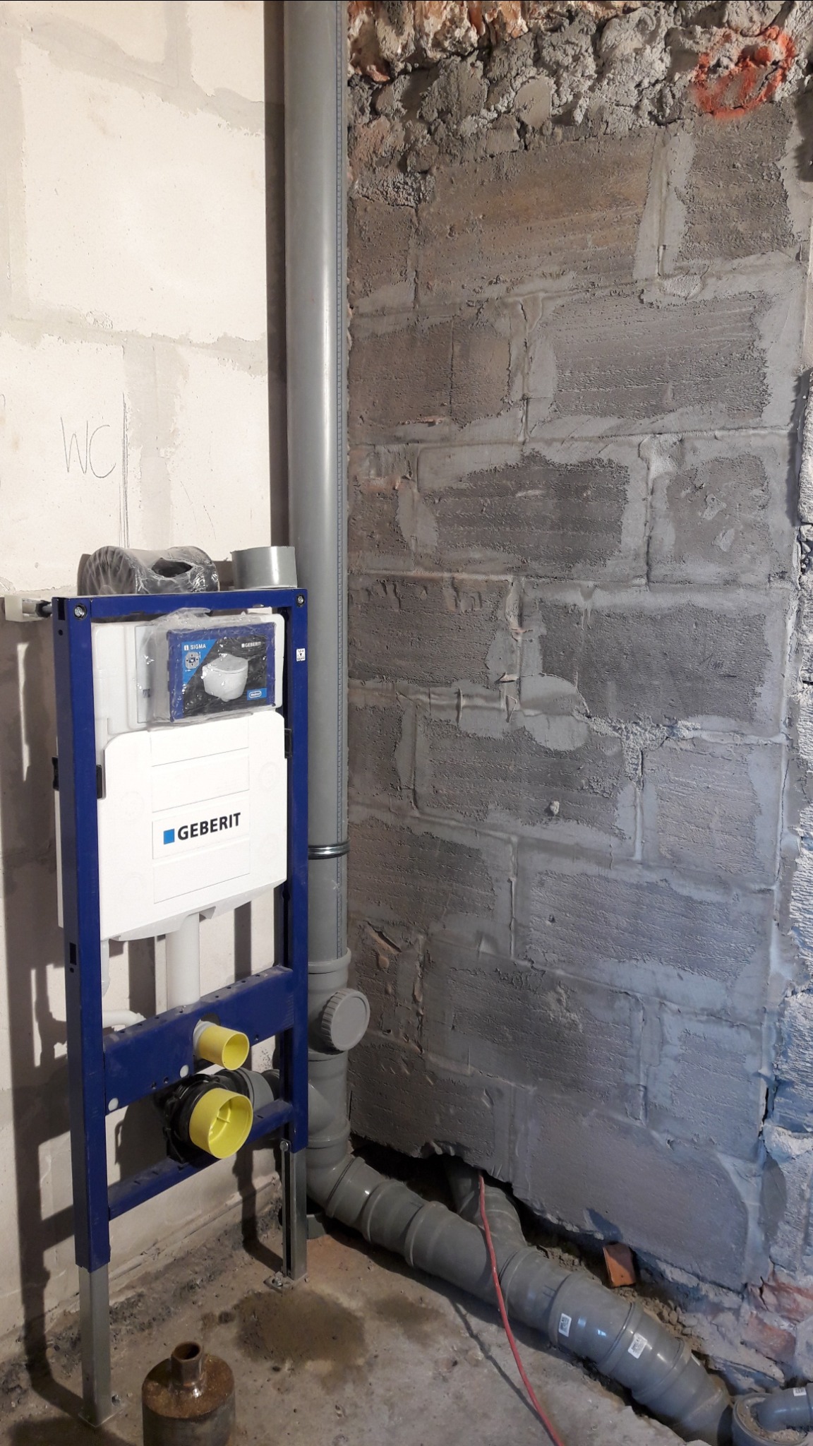 Stelaż podtynkowy marki Geberit zamontowany w surowej łazience, widoczne rury kanalizacyjne i fragment ściany z cegieł.