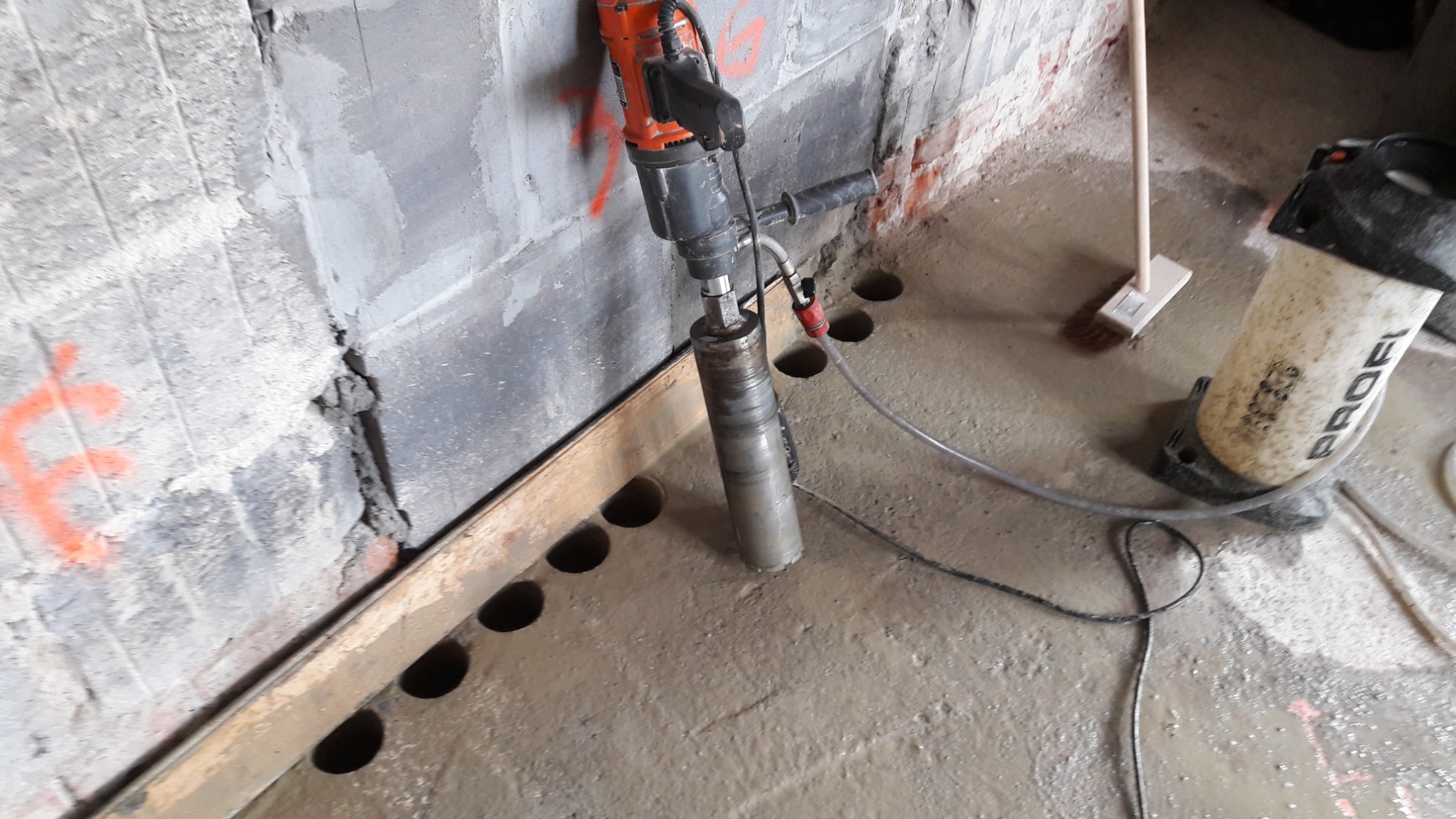 Wiertnica rdzeniowa wiercąca otwory w mokrej posadzce pod ogrzewanie podłogowe, widoczny brudny spryskiwacz i szczotka do zamiatania.
