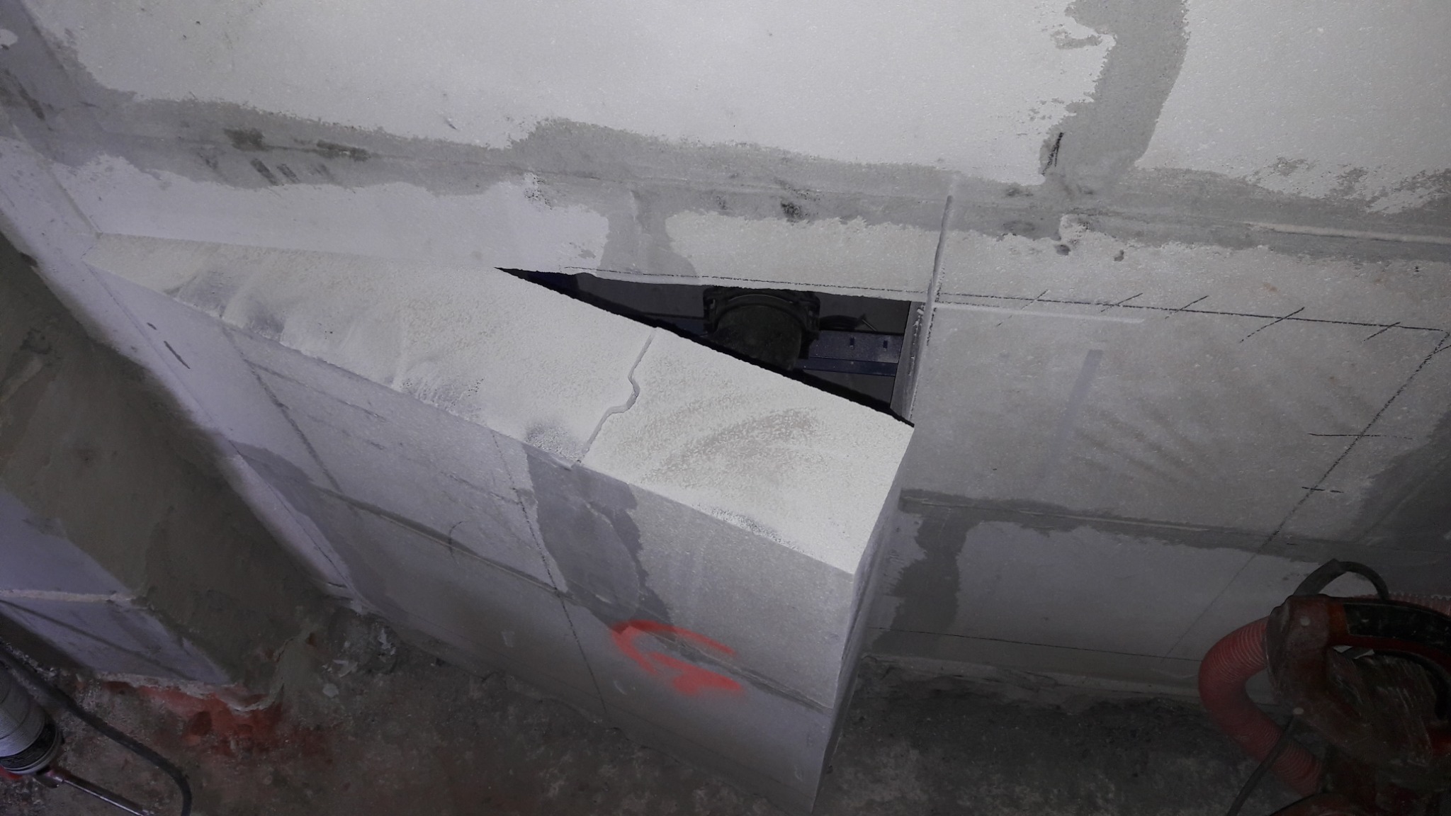 Szara, surowa konstrukcja z betonu komórkowego, widoczne zarysowane linie pomiarowe na suficie i ścianach, fragment otworu z widoczną instalacją wentylacyjną, czerwone oznaczenia na ścianie, w tle...