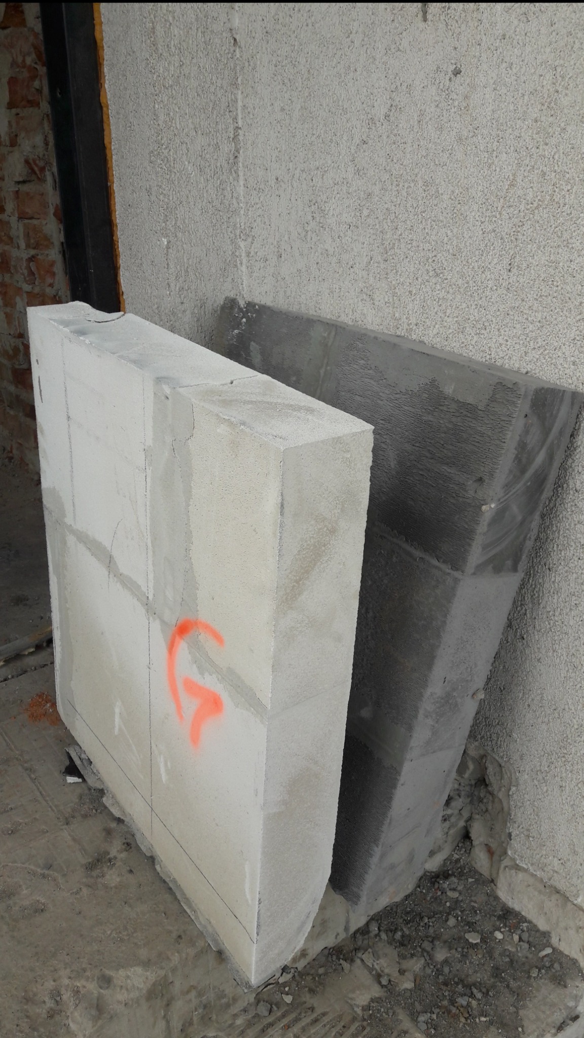 Dwa bloki betonowe, jeden biały z pomarańczowym oznaczeniem, drugi szary, ustawione przy szorstkiej ścianie w trakcie prac budowlanych.
